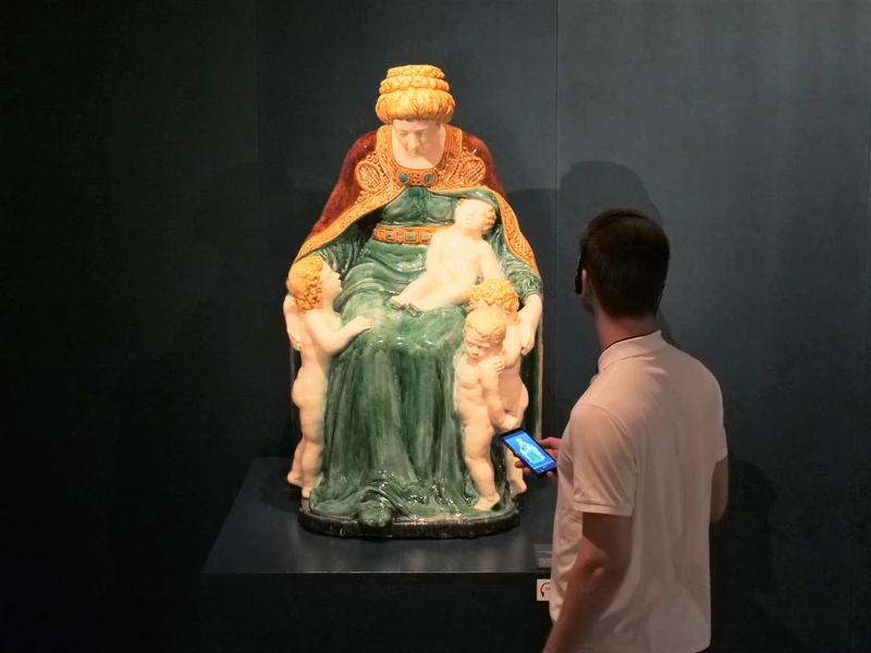 KI generiert: Das Bild zeigt eine Skulptur einer sitzenden Frau mit drei Kindern, die sie umgeben und berühren. Ein Mann steht vor der Skulptur und betrachtet sie, während er ein Audioguide-Gerät in der Hand hält.