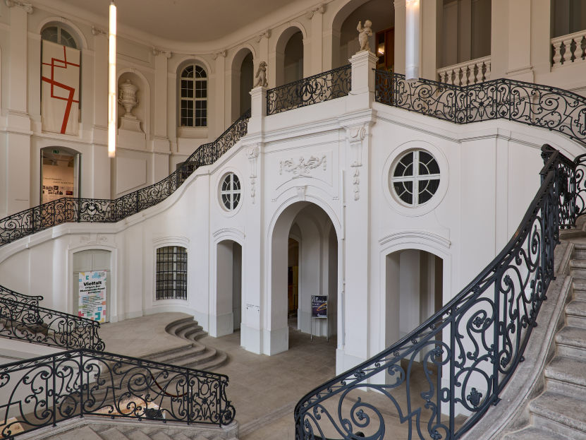 KI generiert: Das Bild zeigt ein prächtiges Treppenhaus mit kunstvollen Eisenarbeiten und weißen Wänden in einem historischen Gebäude. Im Hintergrund sind hohe Fenster und eine moderne Kunstinstallation zu sehen.
