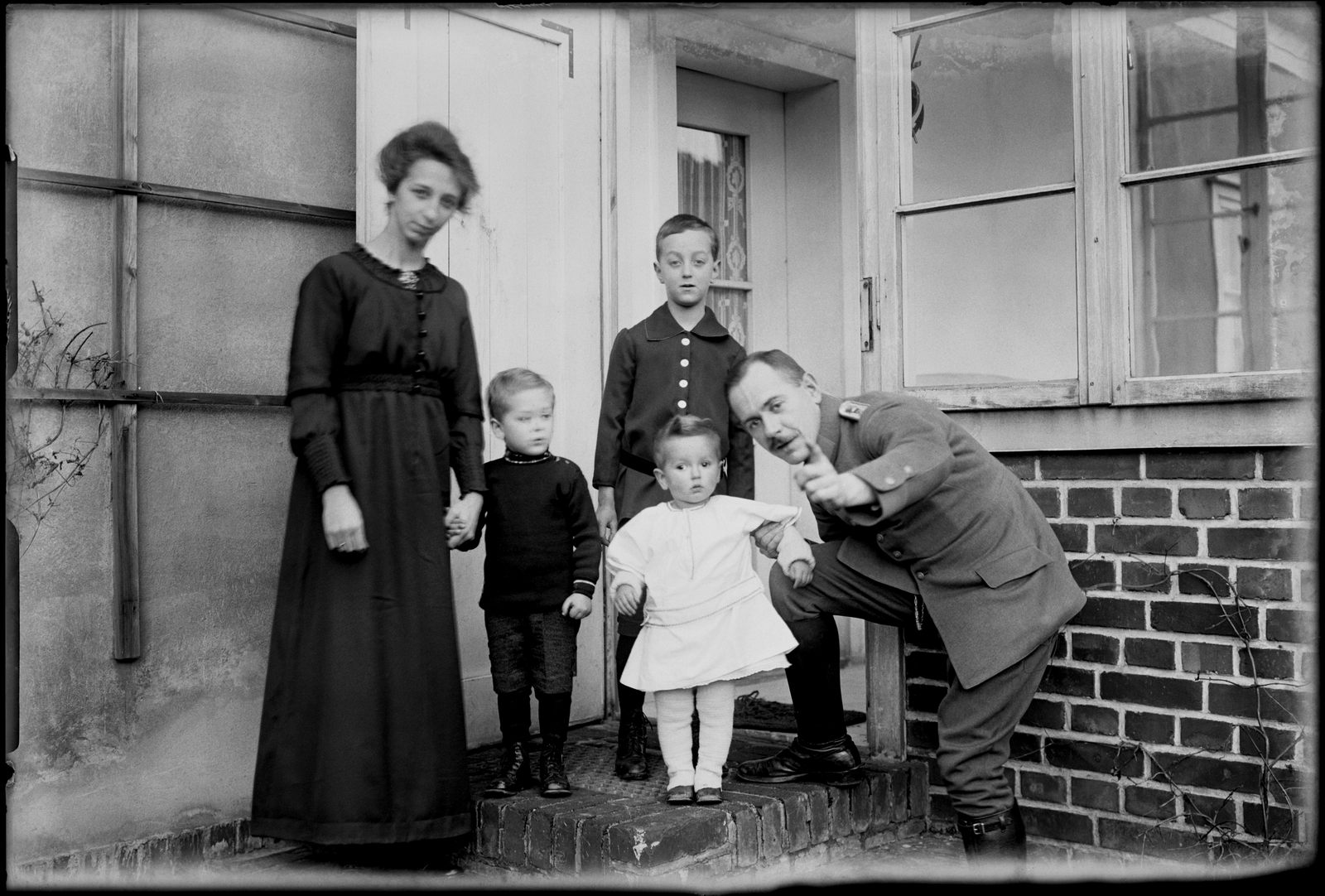 KI generiert: Das Bild zeigt eine Familie, bestehend aus zwei Erwachsenen und drei Kindern, die auf einer Treppe vor einem Gebäude posiert. Ein Mann in Uniform beugt sich fröhlich nach vorne.