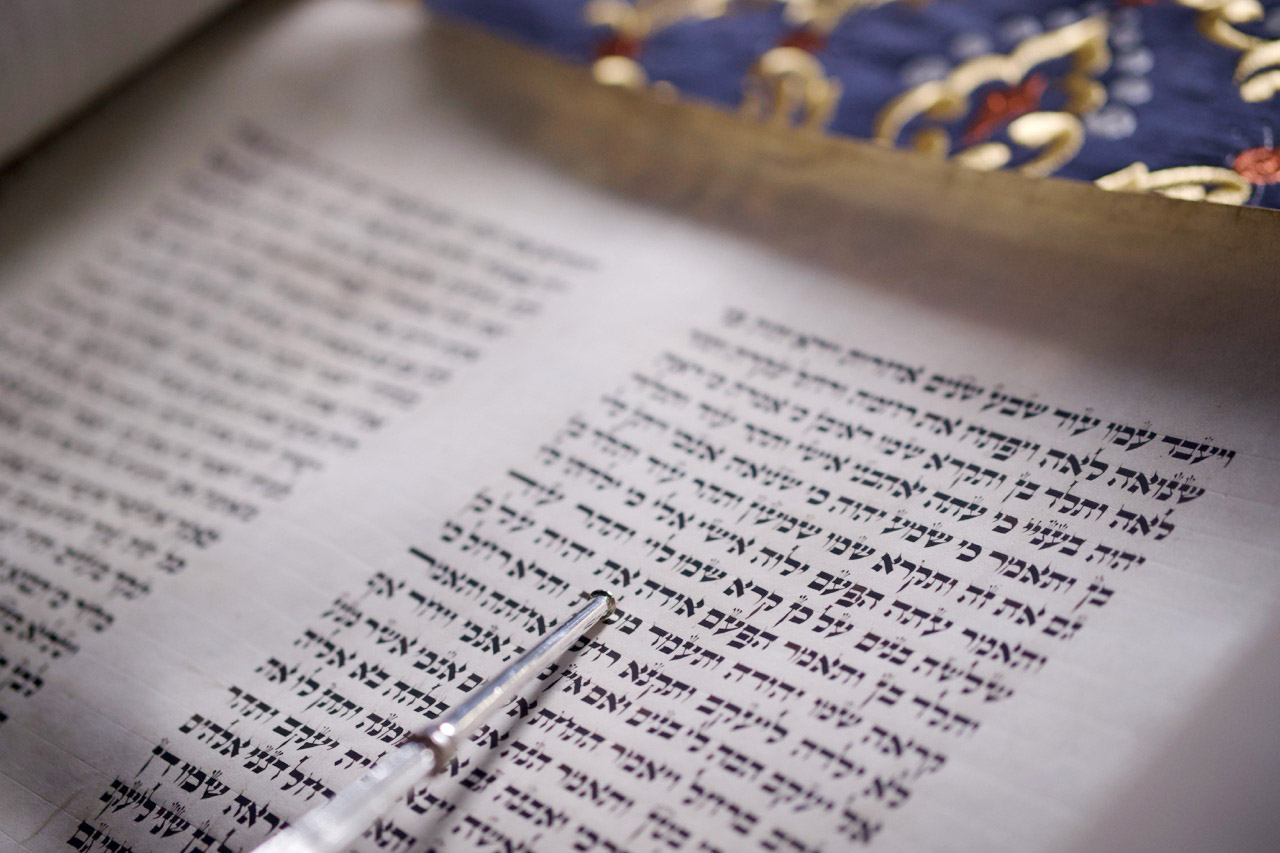 KI generiert: Das Bild zeigt einen Abschnitt einer hebräischen Schriftrolle, bei der ein silberner Zeiger auf die Schrift deutet. Im Hintergrund ist ein bunt verziertes Tuch sichtbar.