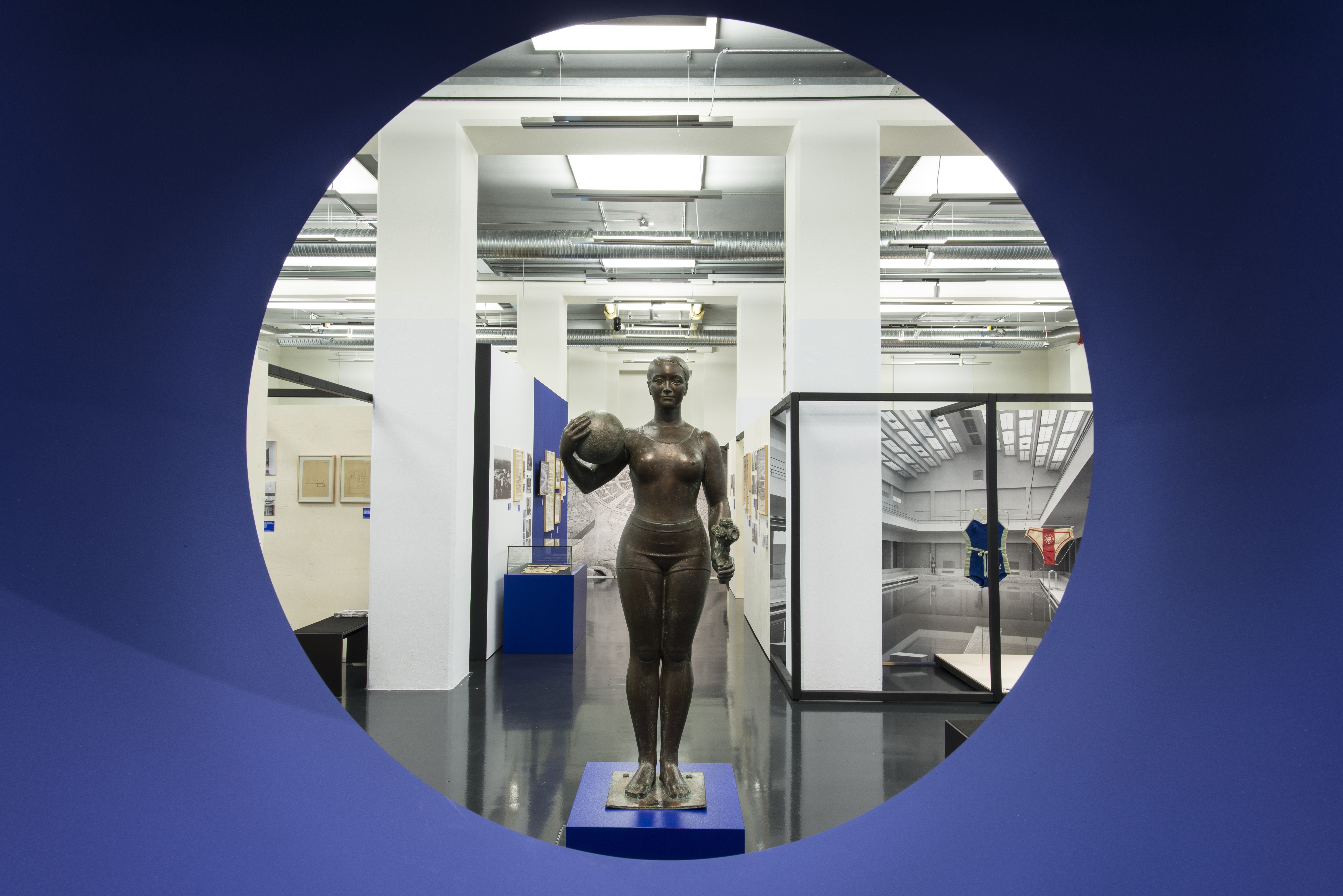 KI generiert: Das Bild zeigt eine Skulptur einer stehenden Frau mit einem Ball in der Hand, ausgestellt in einem modernen Museumsraum. Die Skulptur wird durch eine kreisförmige Öffnung im Vordergrund betrachtet.