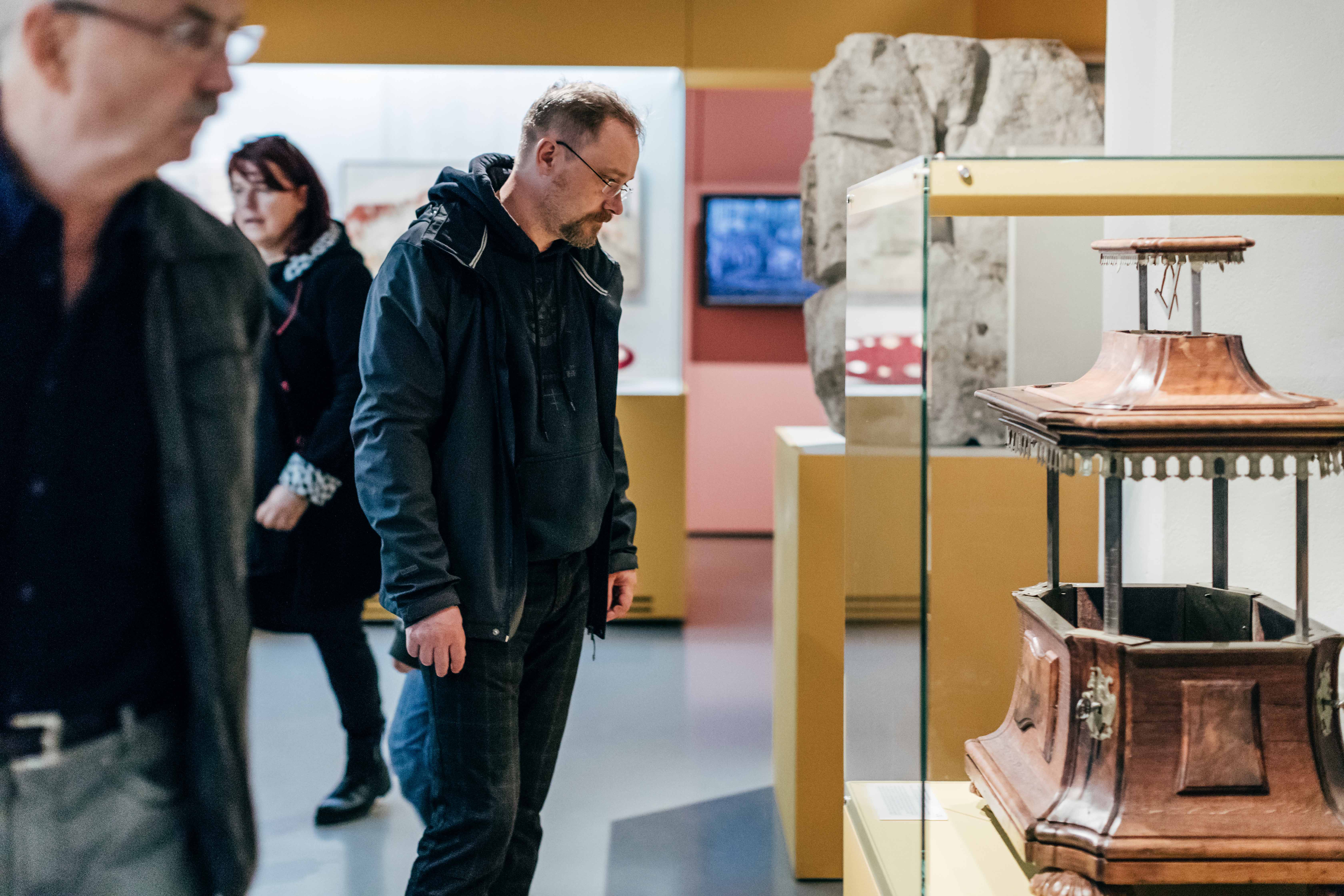 KI generiert: Das Bild zeigt mehrere Menschen, die ein Museum besuchen und sich die Ausstellungsstücke ansehen. Im Vordergrund betrachtet ein Mann ein Artefakt in einer Vitrine.