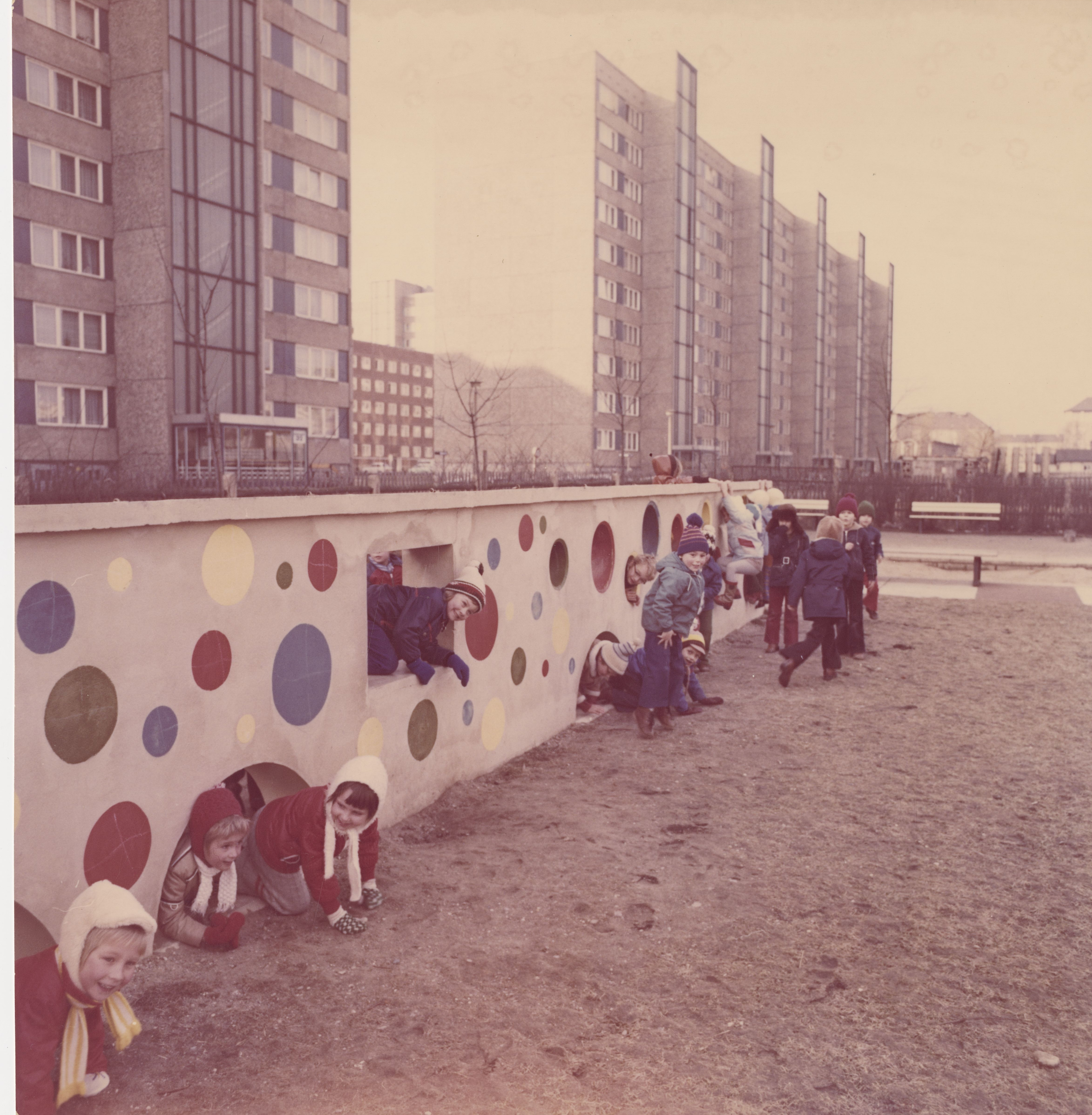 AI generated: The image shows a group of children playing in a playground and crawling and climbing through a colourfully painted wall with circles. Large residential buildings can be seen in the background.