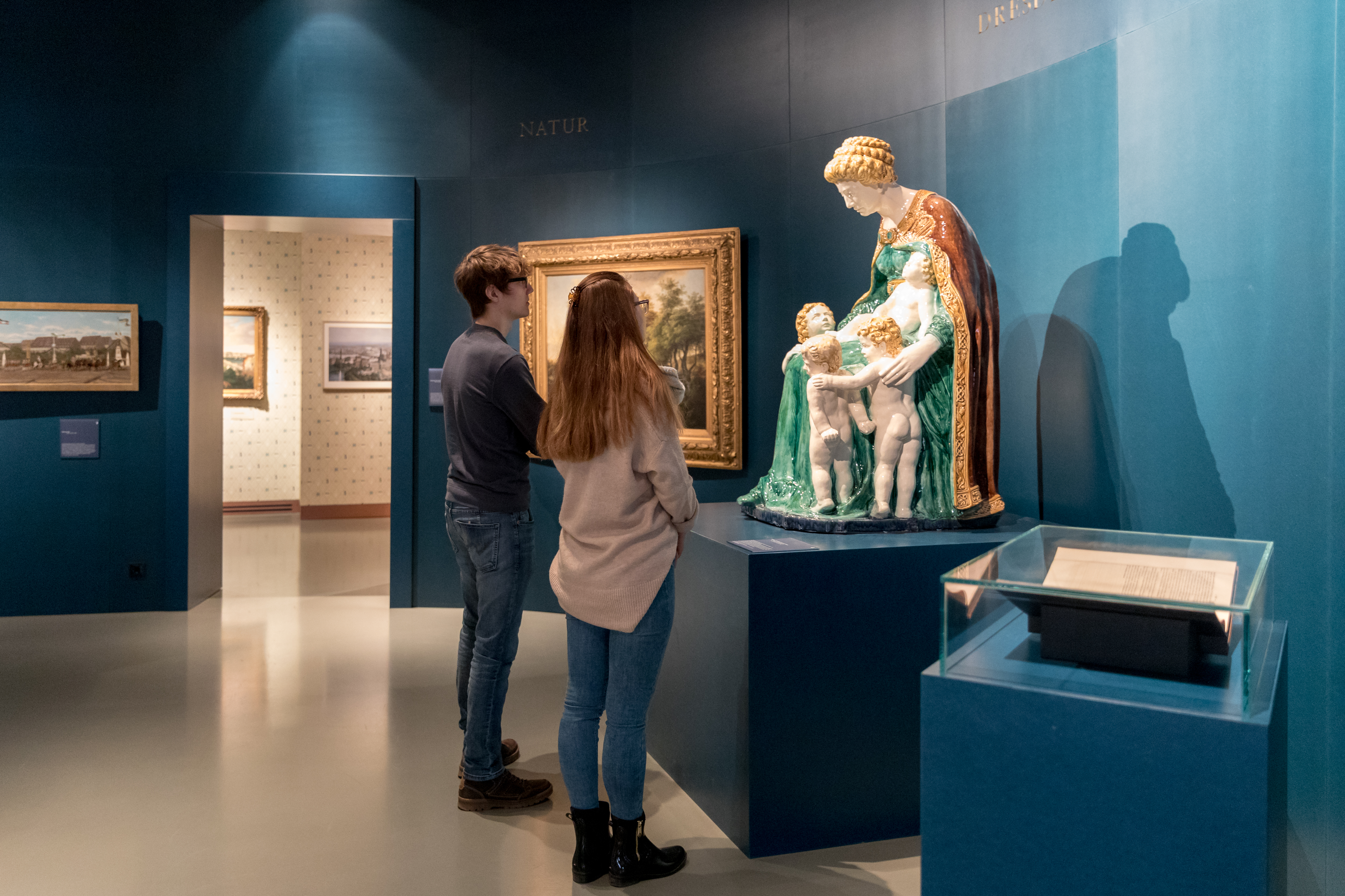 KI generiert: Zwei Personen betrachten in einem Museum eine Skulptur, die eine Frau mit drei Kindern darstellt. Im Hintergrund sind Gemälde an den Wänden zu sehen.