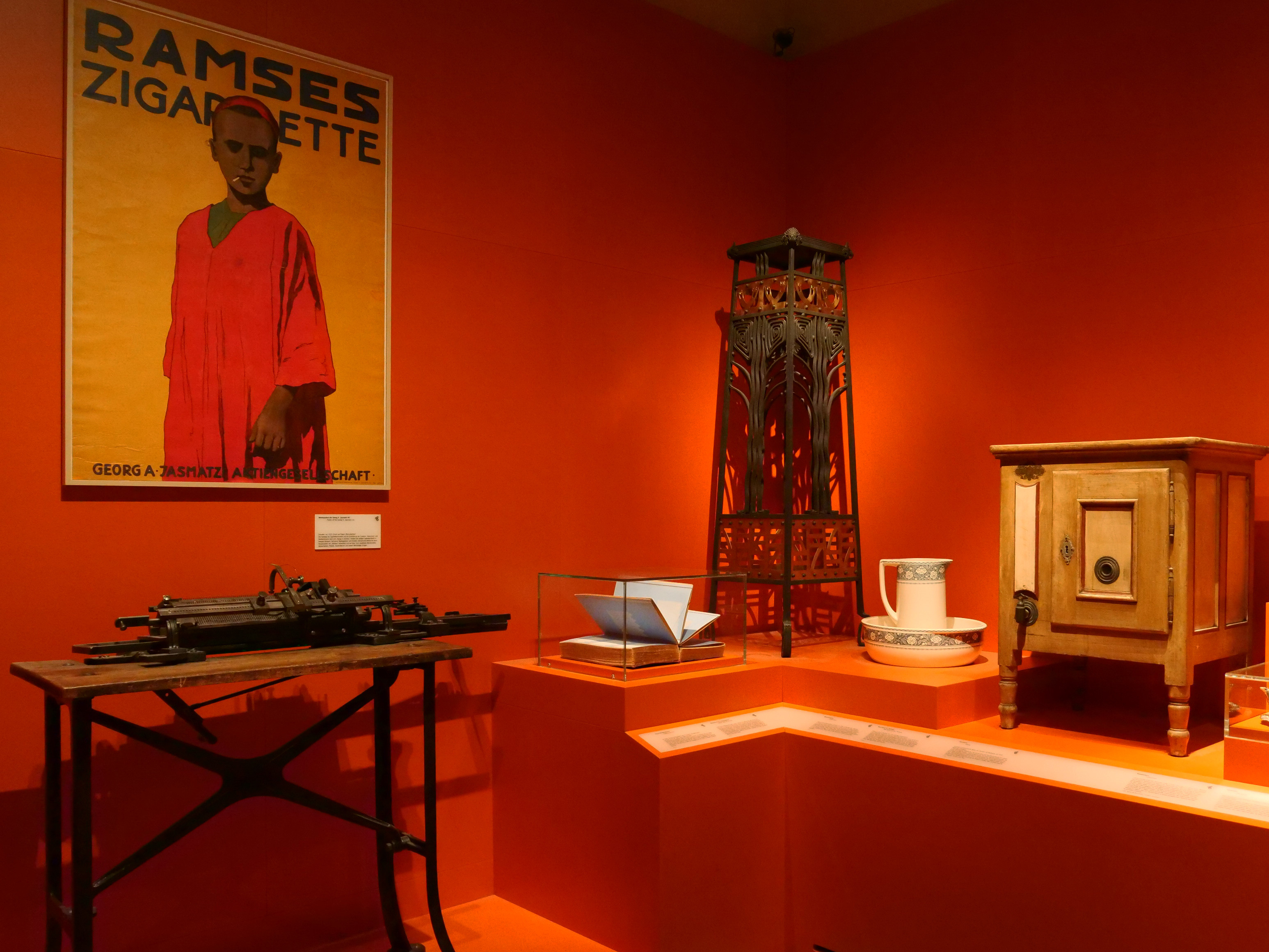 KI generiert: Das Bild zeigt eine Ausstellung mit vintage Objekten, darunter ein Werbeplakat für Zigaretten, ein Safe und verschiedene Designerstücke auf orangefarbenem Hintergrund. Die Exponate scheinen aus der Art-déco-Zeit zu stammen.