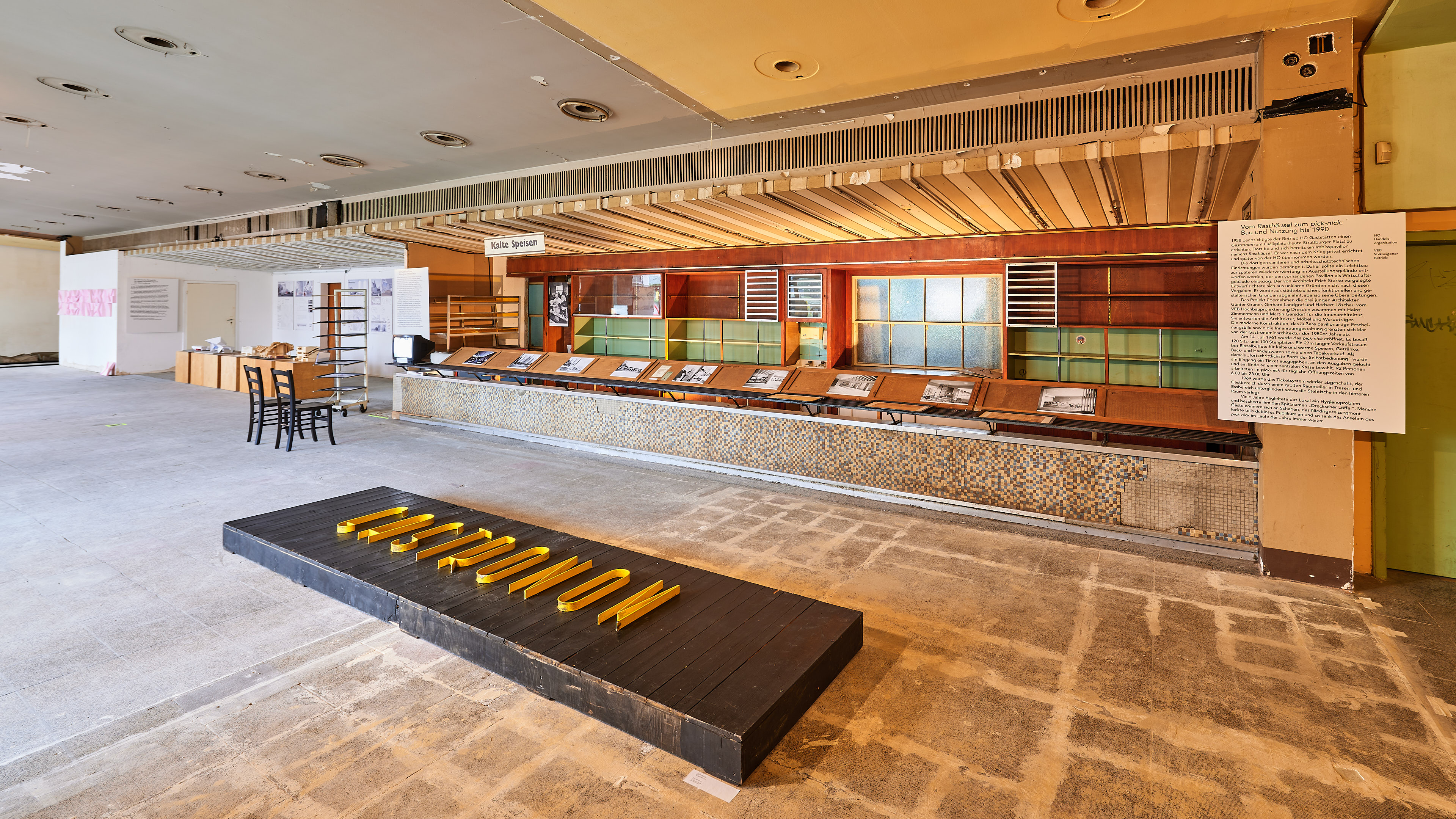 KI generiert: Das Bild zeigt einen verlassenen Raum, der scheinbar als Ausstellungsfläche genutzt wird, mit einer langen Theke, auf der Informationen ausgestellt sind. Auf dem Boden befindet sich eine Plattform mit dem Wort "RESTRIKTION" aus gelben Buchstaben.