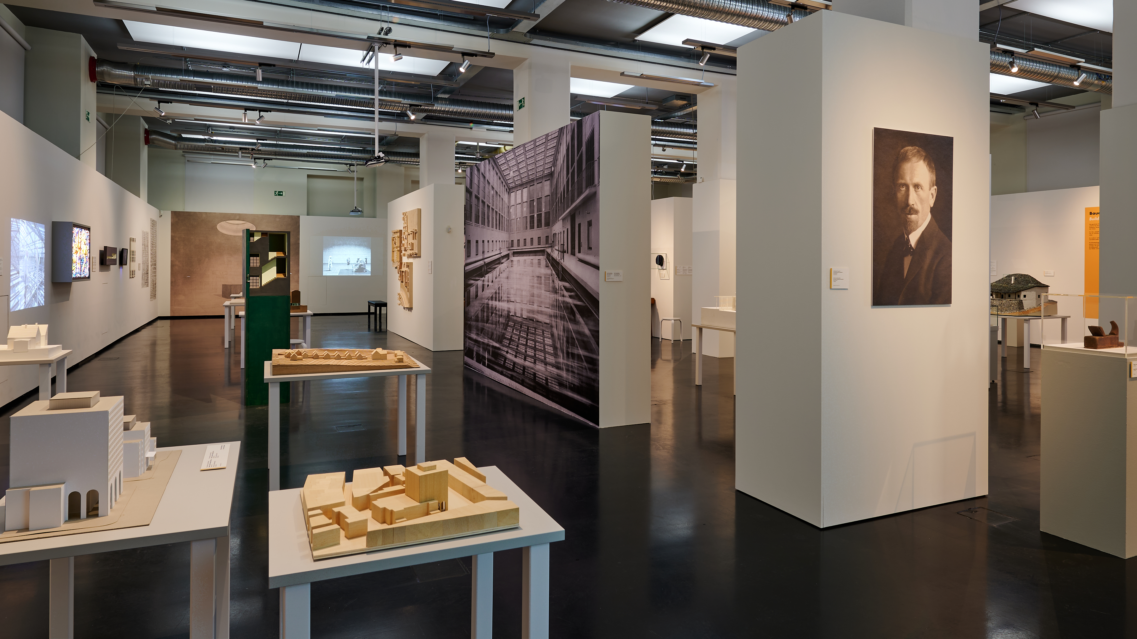 KI generiert: Das Bild zeigt eine Ausstellungshalle mit architektonischen Modellen und Fotografien. Der Raum ist klar strukturiert und gut beleuchtet, was eine moderne und sachliche Atmosphäre schafft.