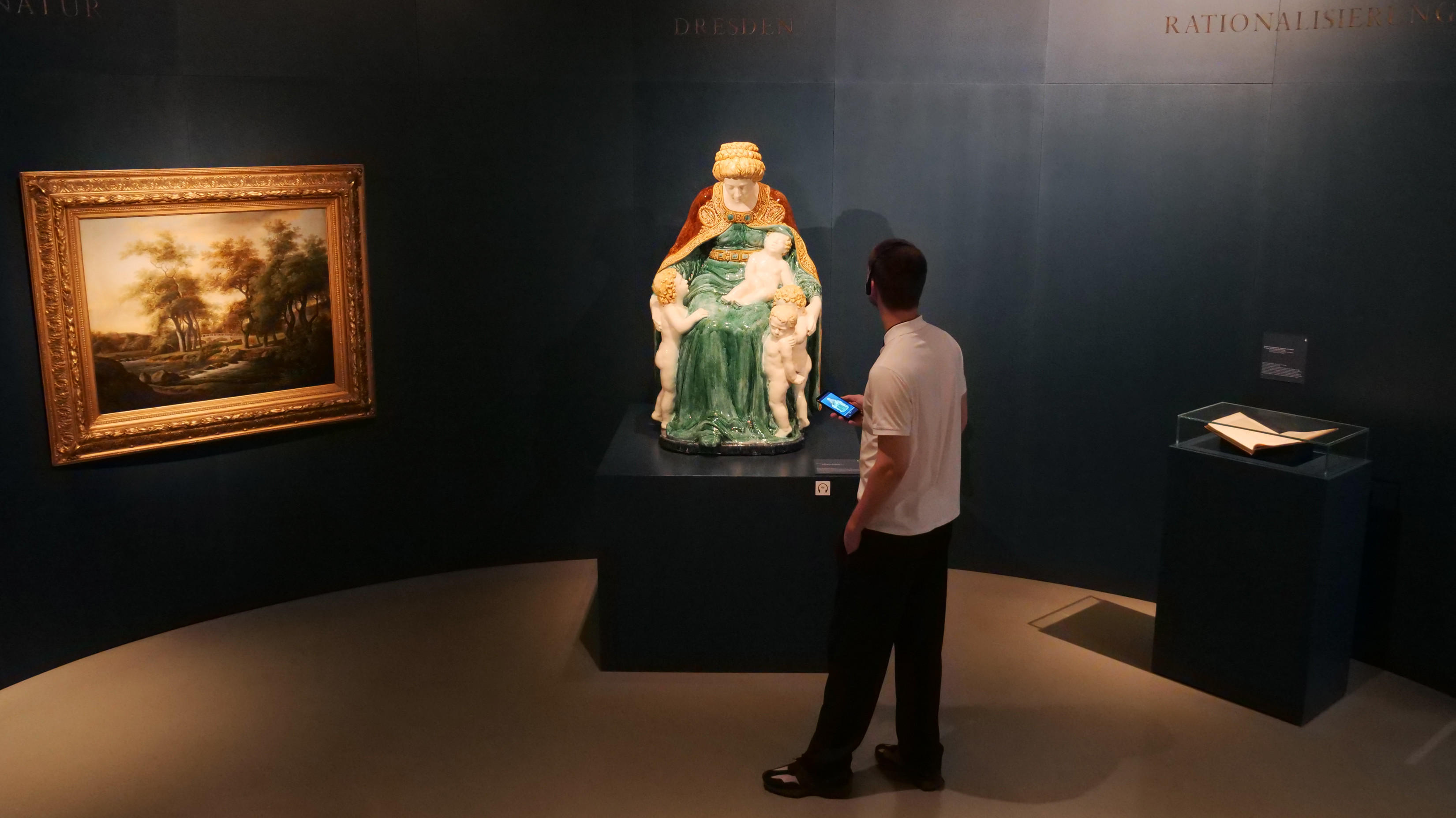 KI generiert: Ein Mann betrachtet in einem Museumsraum eine farbenfrohe Skulptur, die auf einem Podest ausgestellt ist. An der Wand hängt ein gerahmtes Gemälde.