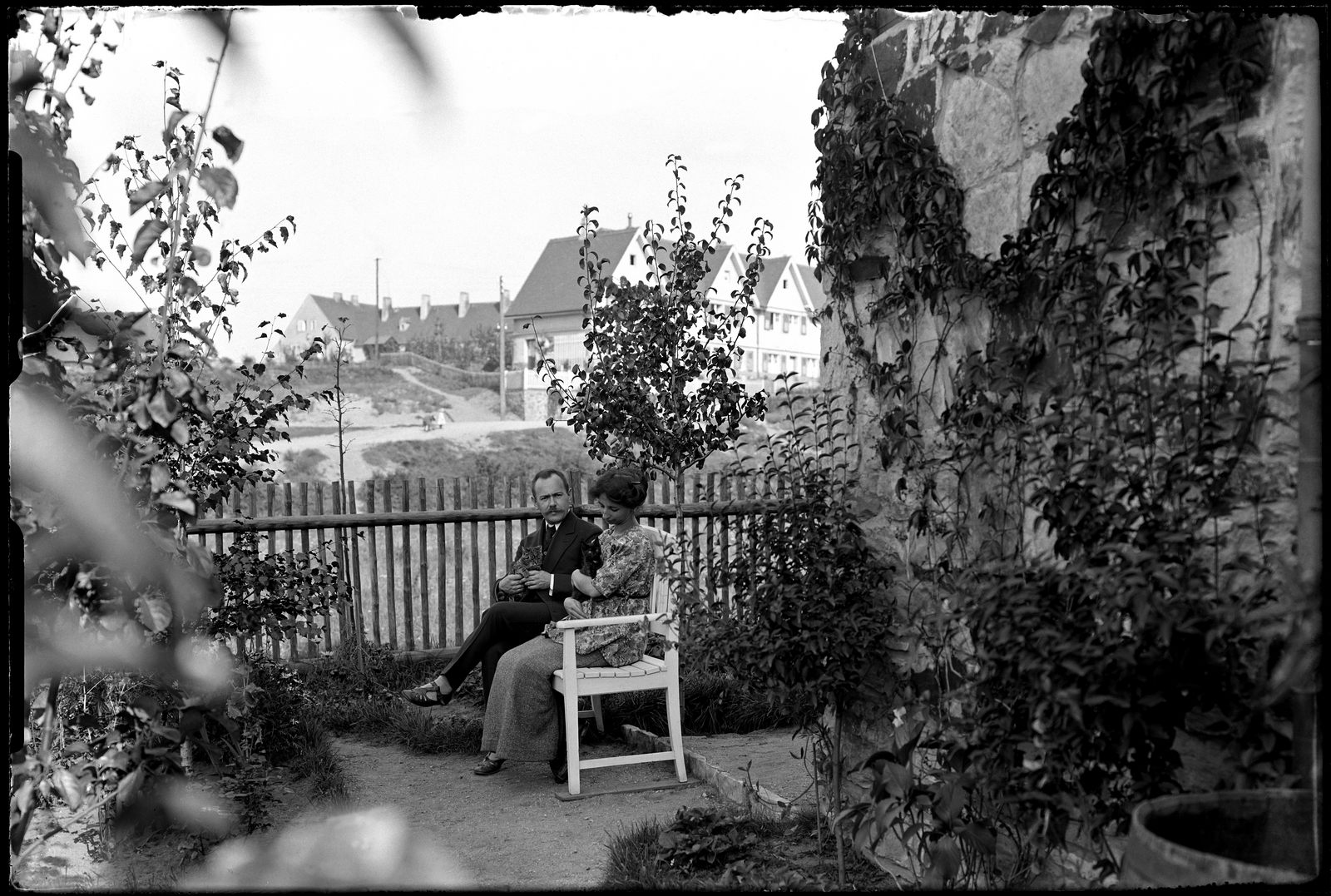 KI generiert: Ein Paar sitzt in einem Garten auf einer Bank, umgeben von Pflanzen und Sträuchern. Im Hintergrund sind Häuser und ein Gartenzaun zu sehen.