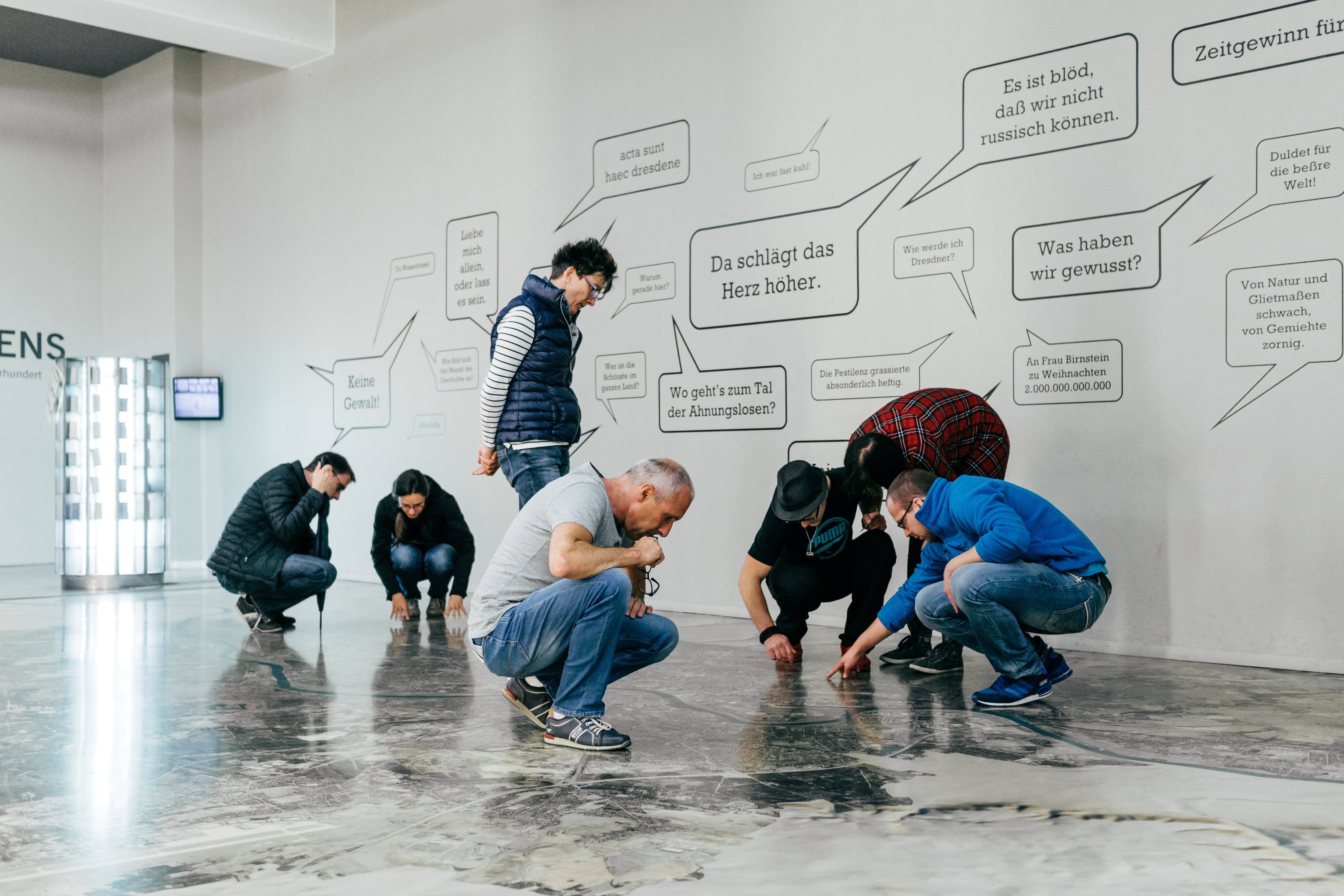 KI generiert: Mehrere Personen sind in einem offenen Raum und inspizieren oder betrachten den Boden, während an der Wand hinter ihnen verschiedene Sprechblasen mit deutschen Texten angebracht sind. Die Szene deutet auf eine interaktive oder erzieherische Ausstellung hin.