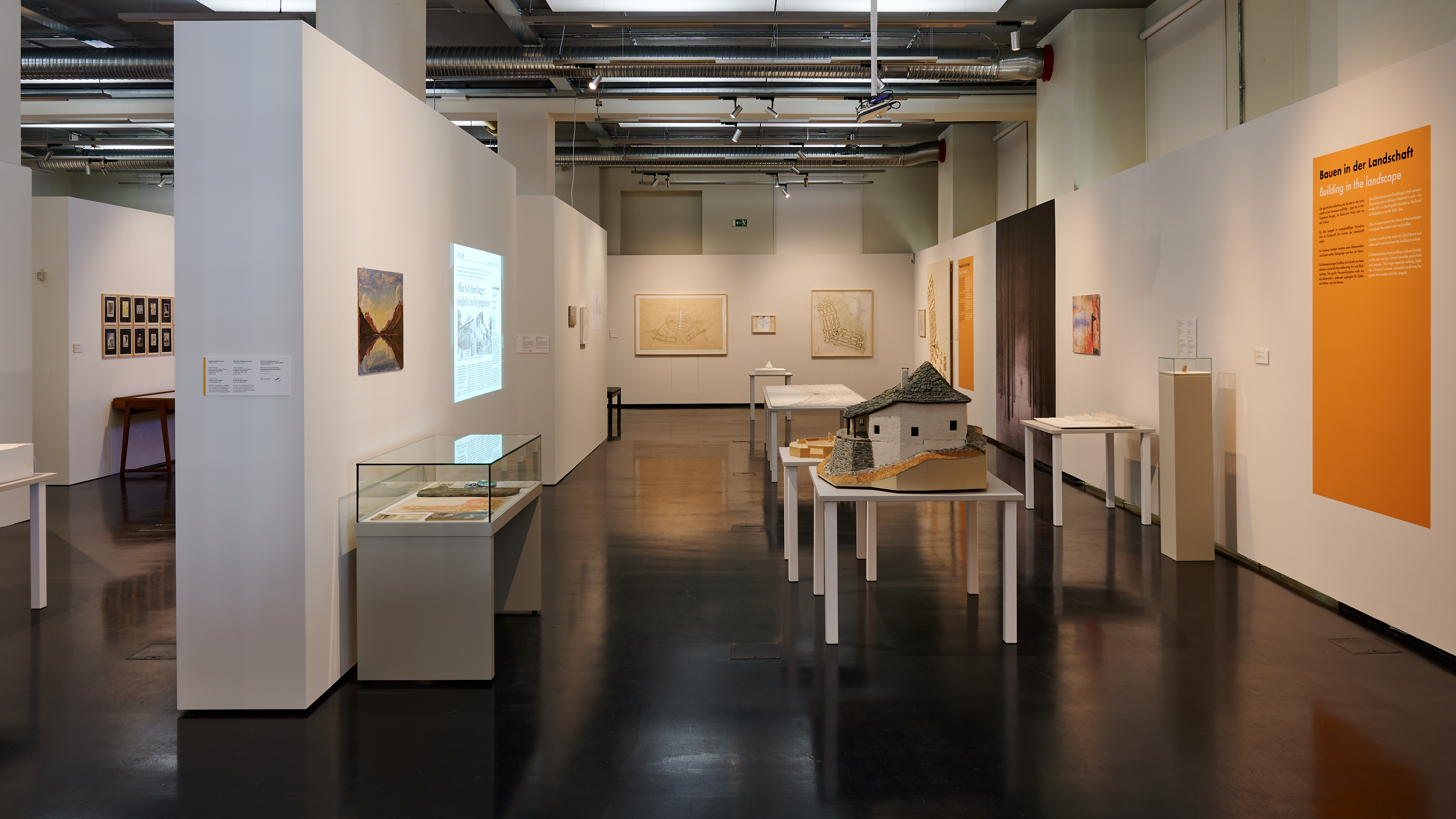 KI generiert: Das Bild zeigt eine Ausstellungsgalerie mit verschiedenen Exponaten, darunter Modelle und Bilder, die auf weißen Tischen und an den Wänden präsentiert werden. Die Umgebung wirkt ordentlich und strukturiert, mit informativen Tafeln an den Wänden.