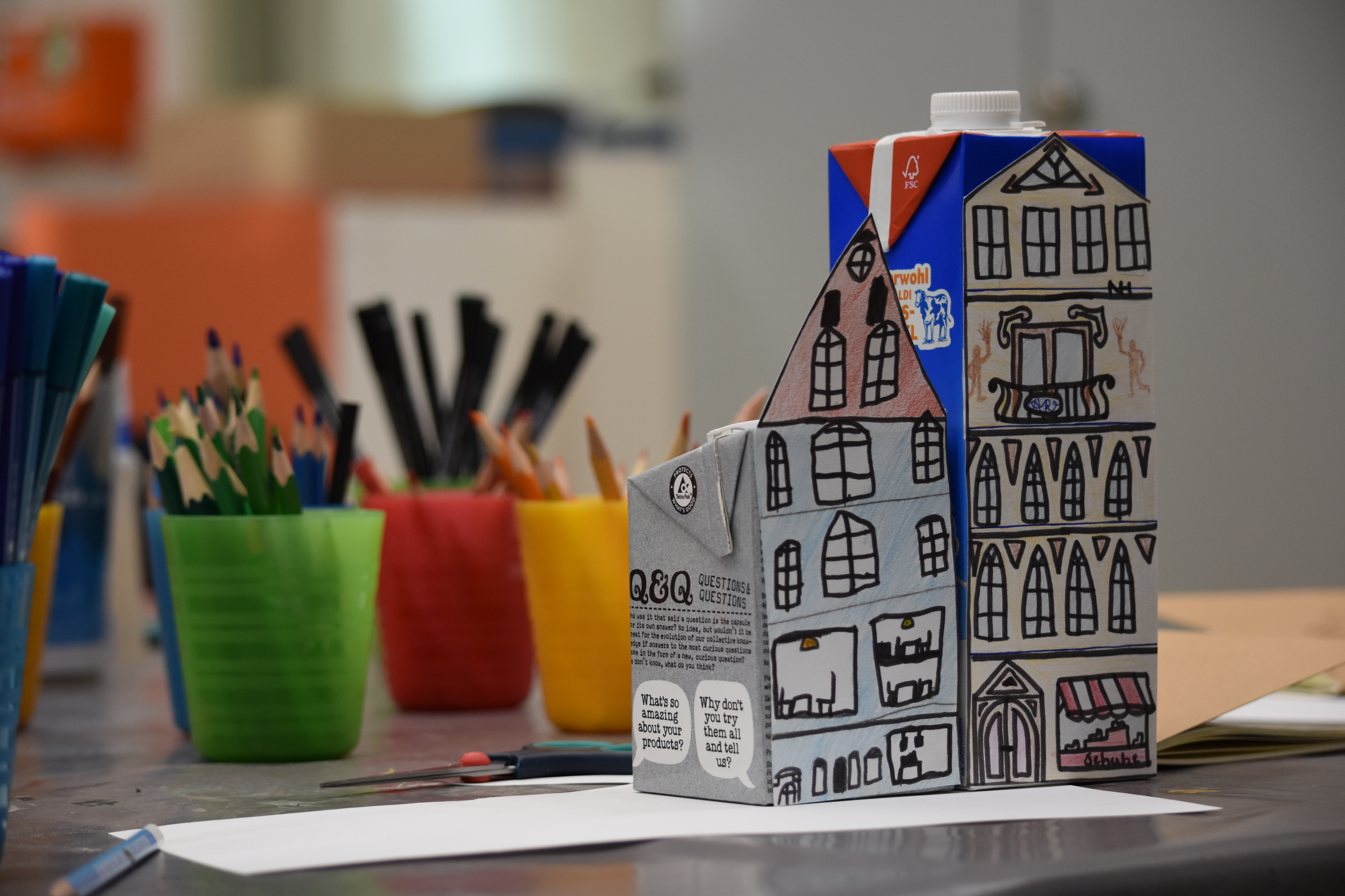 KI generiert: Das Bild zeigt eine Milchtüte, die künstlerisch mit einem Stadthausdesign bemalt wurde. Im Hintergrund sind bunte Behälter mit Buntstiften zu sehen.