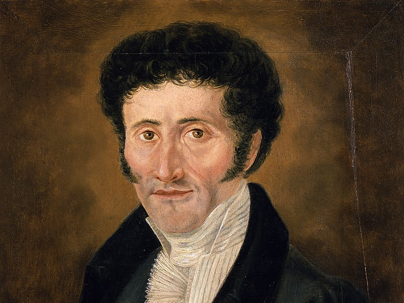 KI generiert: Das Bild zeigt ein Porträt eines Mannes mit dunklen lockigen Haaren und Koteletten, der formelle Kleidung trägt. Der Hintergrund ist dunkel und schlicht gehalten.