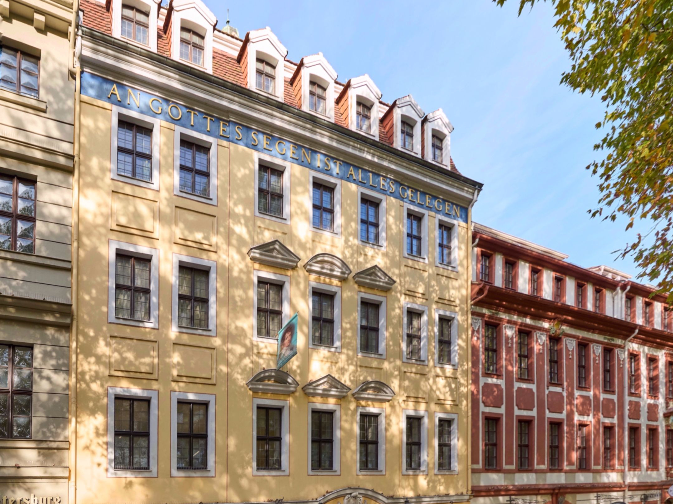 KI generiert: Das Bild zeigt die Fassade eines mehrstöckigen Gebäudes mit der Aufschrift "AN GOTTES SEGEN IST ALLES GELEGEN". Im Vordergrund sind herbstliche Bäume zu sehen, die teilweise Schatten auf die Fassade werfen.