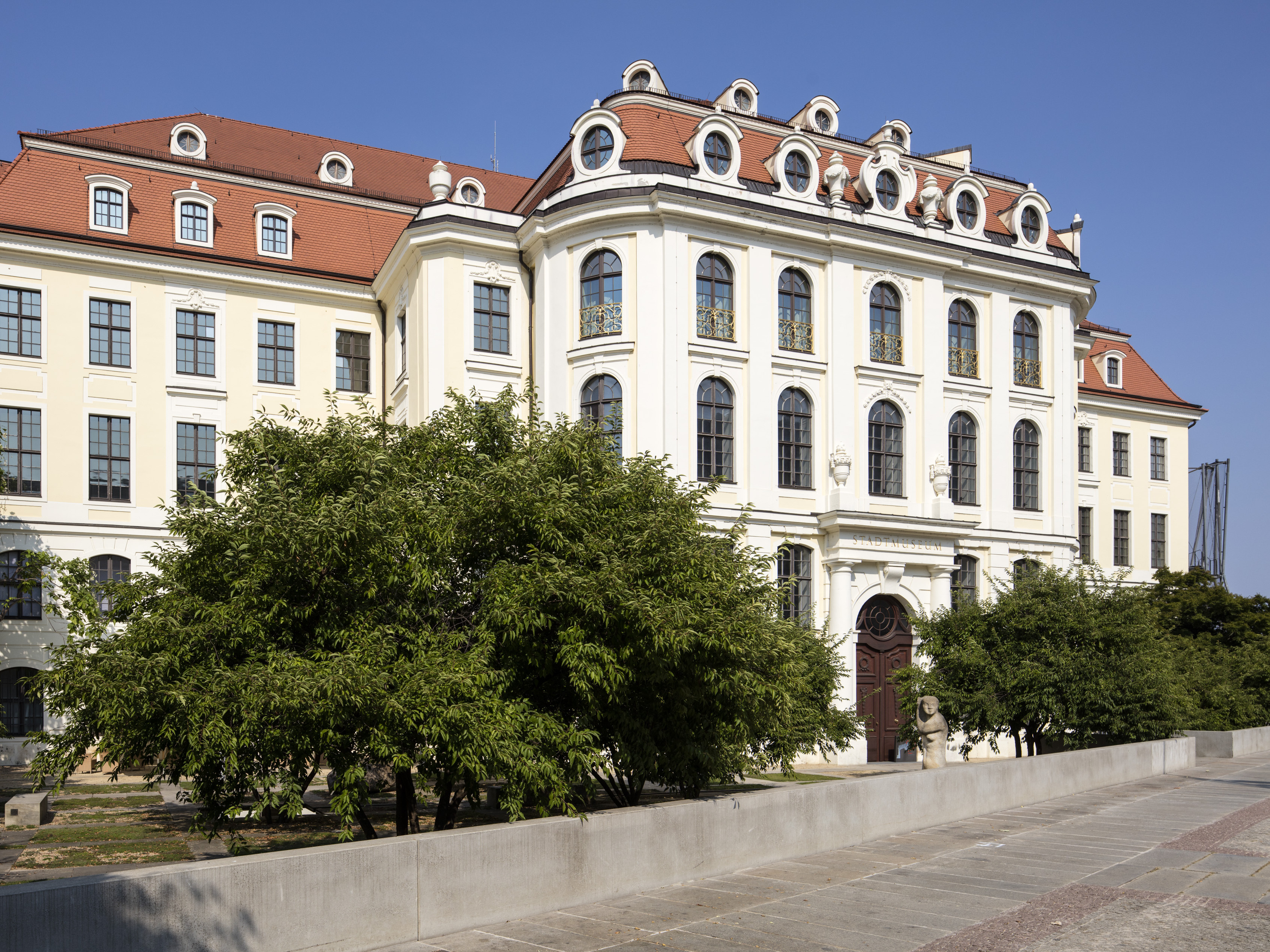KI generiert: Das Bild zeigt ein imposantes historisches Gebäude mit einer reich verzierten Fassade und zahlreichen großen Fenstern. Vor dem Gebäude sind mehrere Bäume und Sträucher zu sehen.
