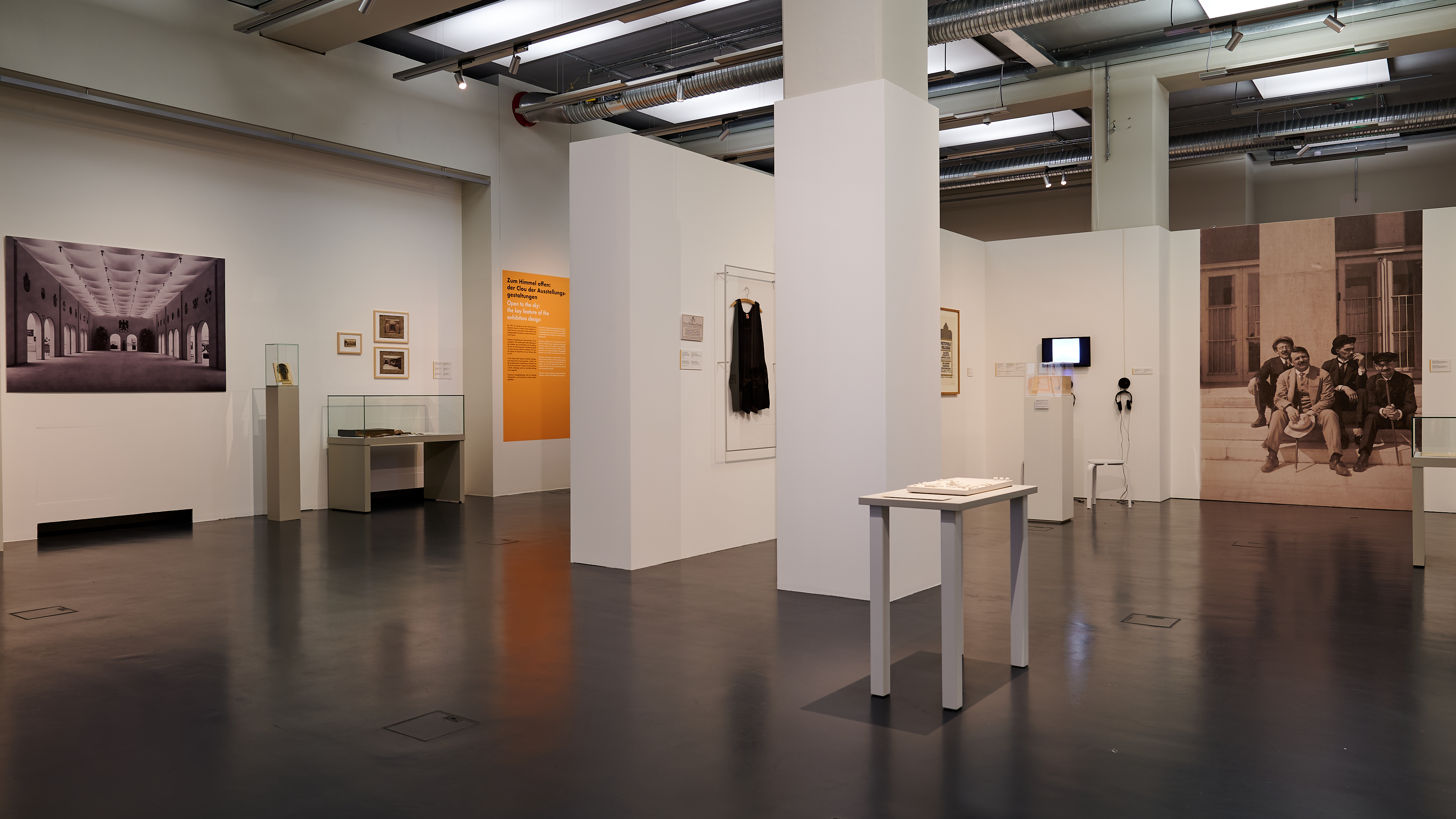 KI generiert: Das Bild zeigt eine Museumsausstellung mit verschiedenen Kunstwerken und Fotografien an den Wänden sowie ausgestellten Kleidungsstücken und Informationsschildern. Der Raum wirkt modern und gut beleuchtet, wobei das Hauptaugenmerk auf den ausgestellten Objekten liegt.