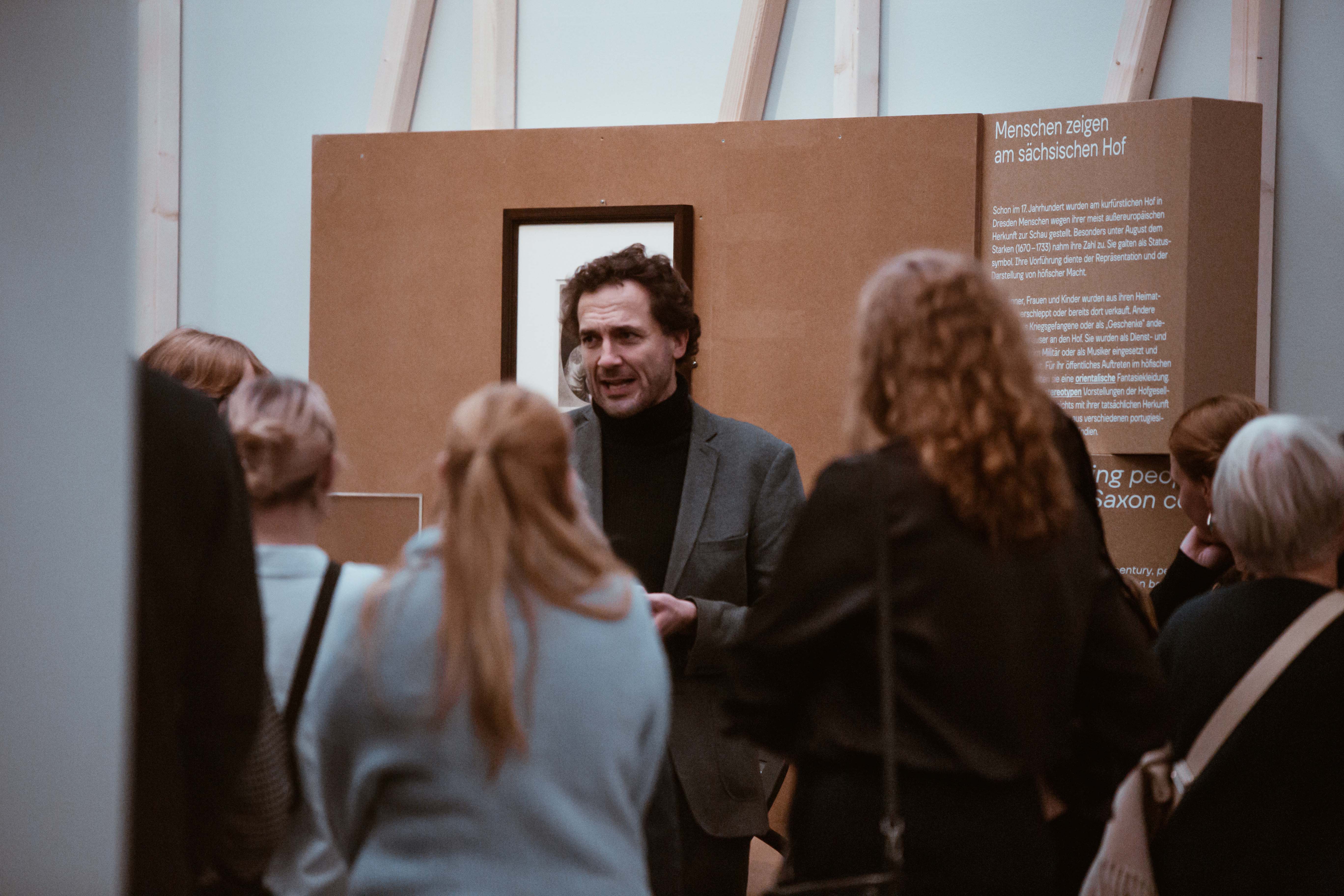 KI generiert: Das Bild zeigt eine Gruppe von Menschen, die anscheinend an einer Führung oder Erklärung in einem Museum oder einer Ausstellung teilnehmen. Ein Mann spricht zu der Gruppe, während die anderen zuhören.