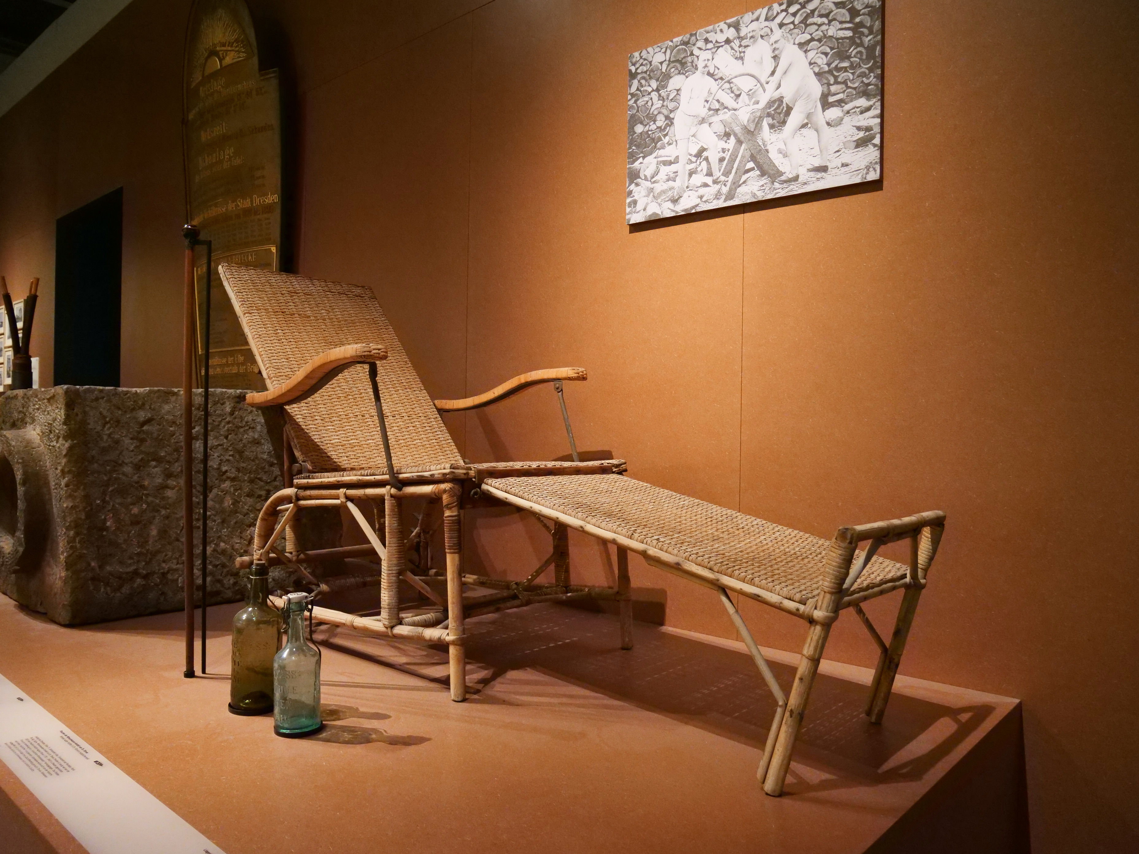 KI generiert: Das Bild zeigt eine Ausstellung mit einem historischen Liegestuhl aus Korbgeflecht und antiken Glasflaschen. Im Hintergrund hängt ein Schwarz-Weiß-Foto, das Menschen beim Arbeiten zeigt.