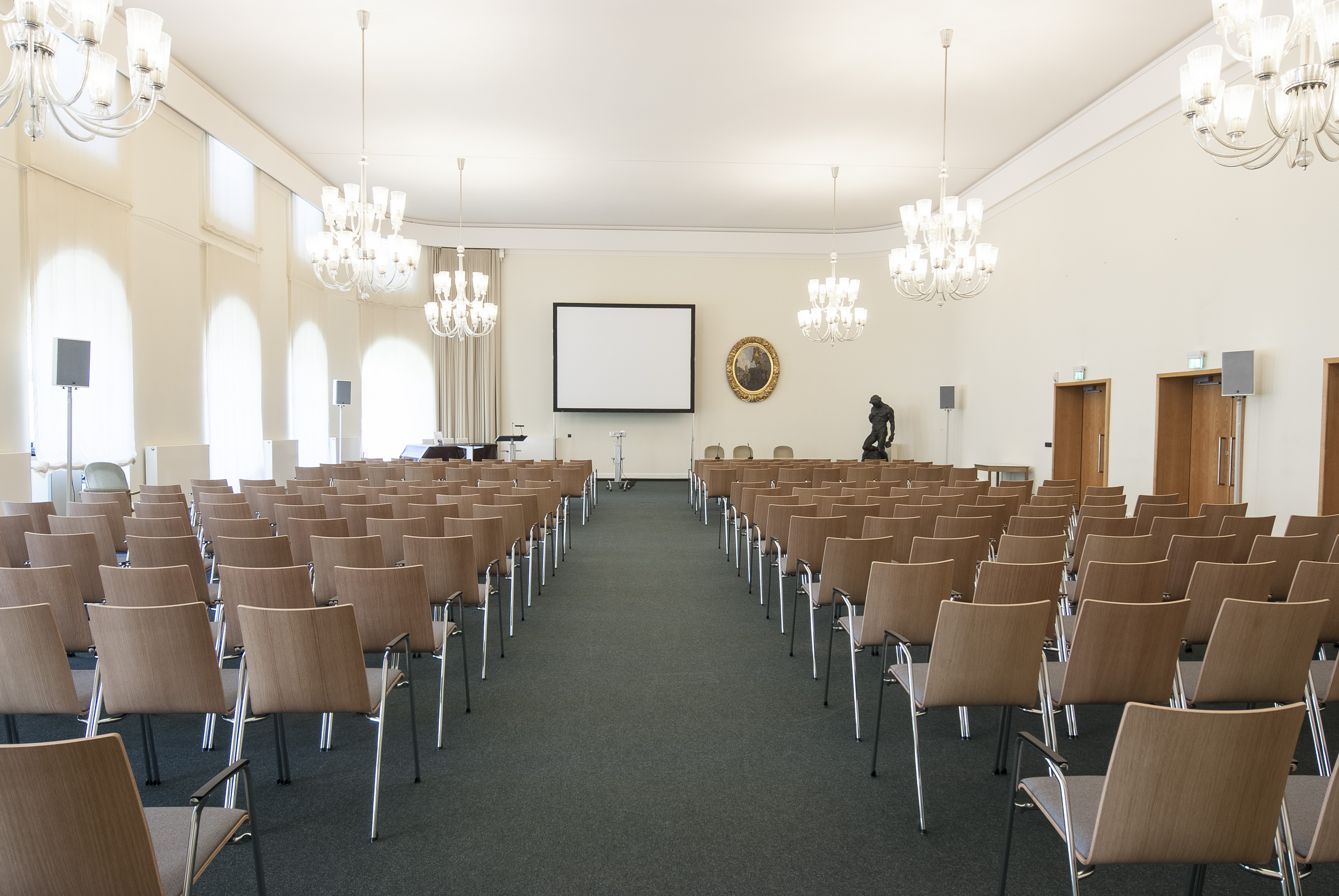 KI generiert: Das Bild zeigt einen leeren Konferenzraum mit Reihen von Stühlen, die auf eine Leinwand ausgerichtet sind. An den Wänden hängen Luster, und im Hintergrund befindet sich ein Podium.