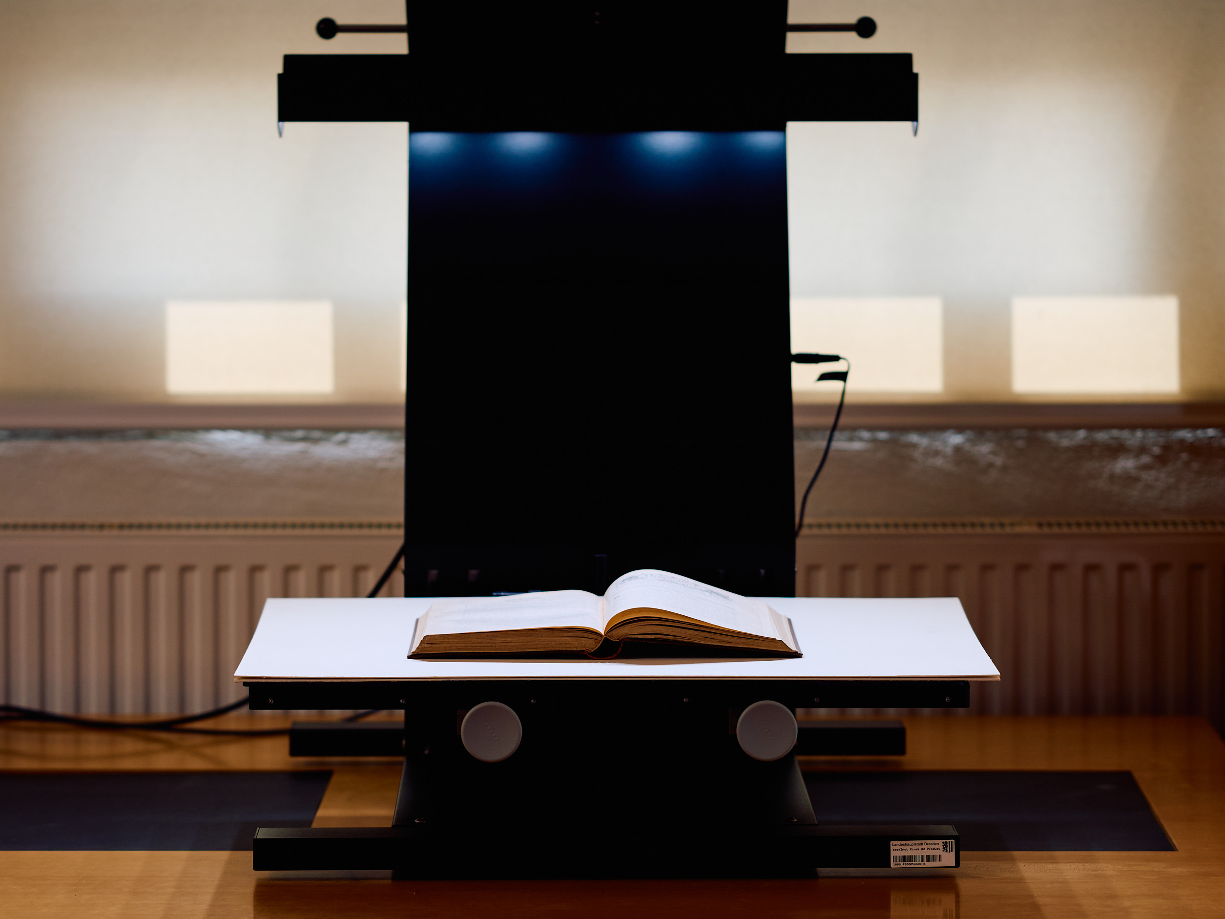 KI generiert: Das Bild zeigt ein aufgeschlagenes Buch auf einem Scanner-Tisch, bereit zur Digitalisierung. Der Scanner ist beleuchtet und steht in einem Raum mit indirekter Beleuchtung.