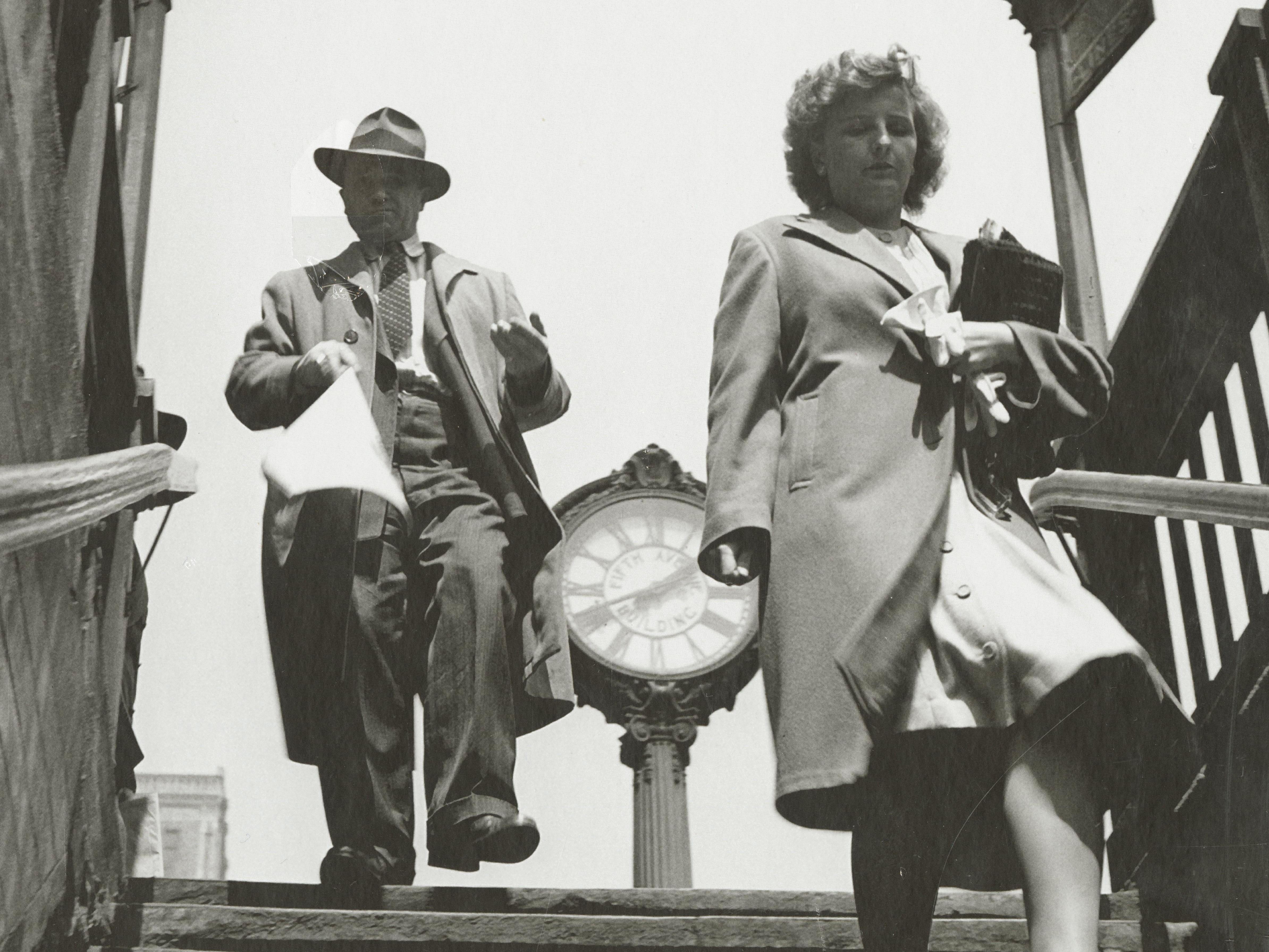 KI generiert: Das Bild zeigt zwei Personen, einen Mann und eine Frau, die eine Treppe hinuntergehen, während im Hintergrund eine große Uhr sichtbar ist. Es handelt sich um eine Schwarz-Weiß-Fotografie mit dem Namen "Fred Stein" im oberen Teil platziert.