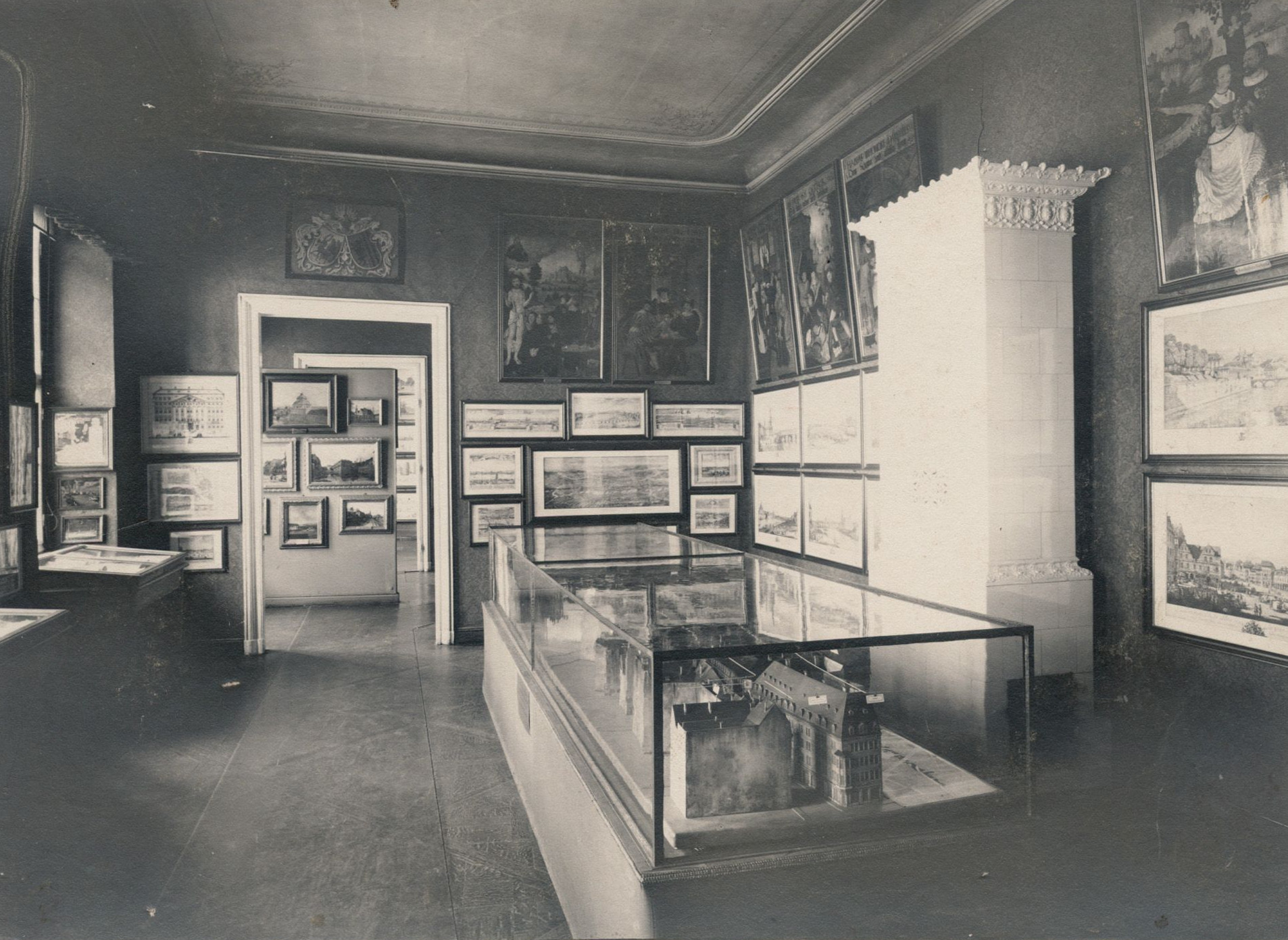 KI generiert: Das Bild zeigt eine Innenansicht eines Raumes im Stadtmuseum Dresden im Jahr 1904. An den Wänden hängen zahlreiche Gemälde und Zeichnungen, während in der Mitte des Raumes Vitrinen mit Exponaten stehen.