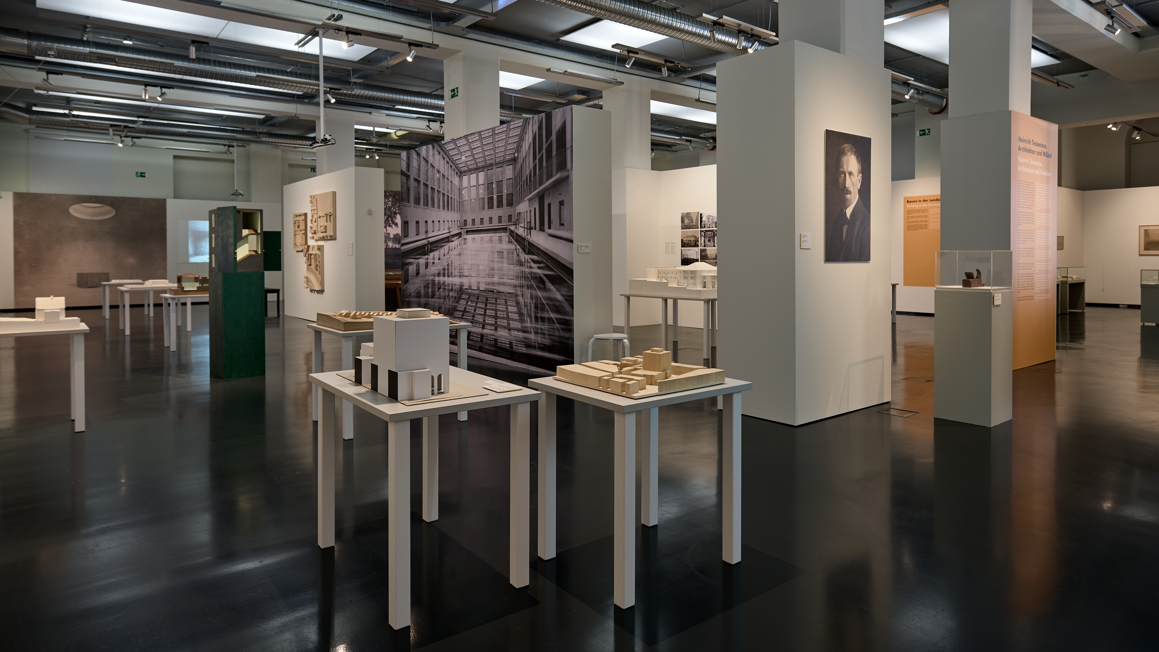 KI generiert: Das Bild zeigt eine Ausstellung in einem modernen Museum oder Galerie mit architektonischen Modellen und großformatigen Fotos an den Wänden. Der Raum ist gut beleuchtet, mit mehreren Tischen und Wänden, die verschiedene architektonische Projekte präsentieren.