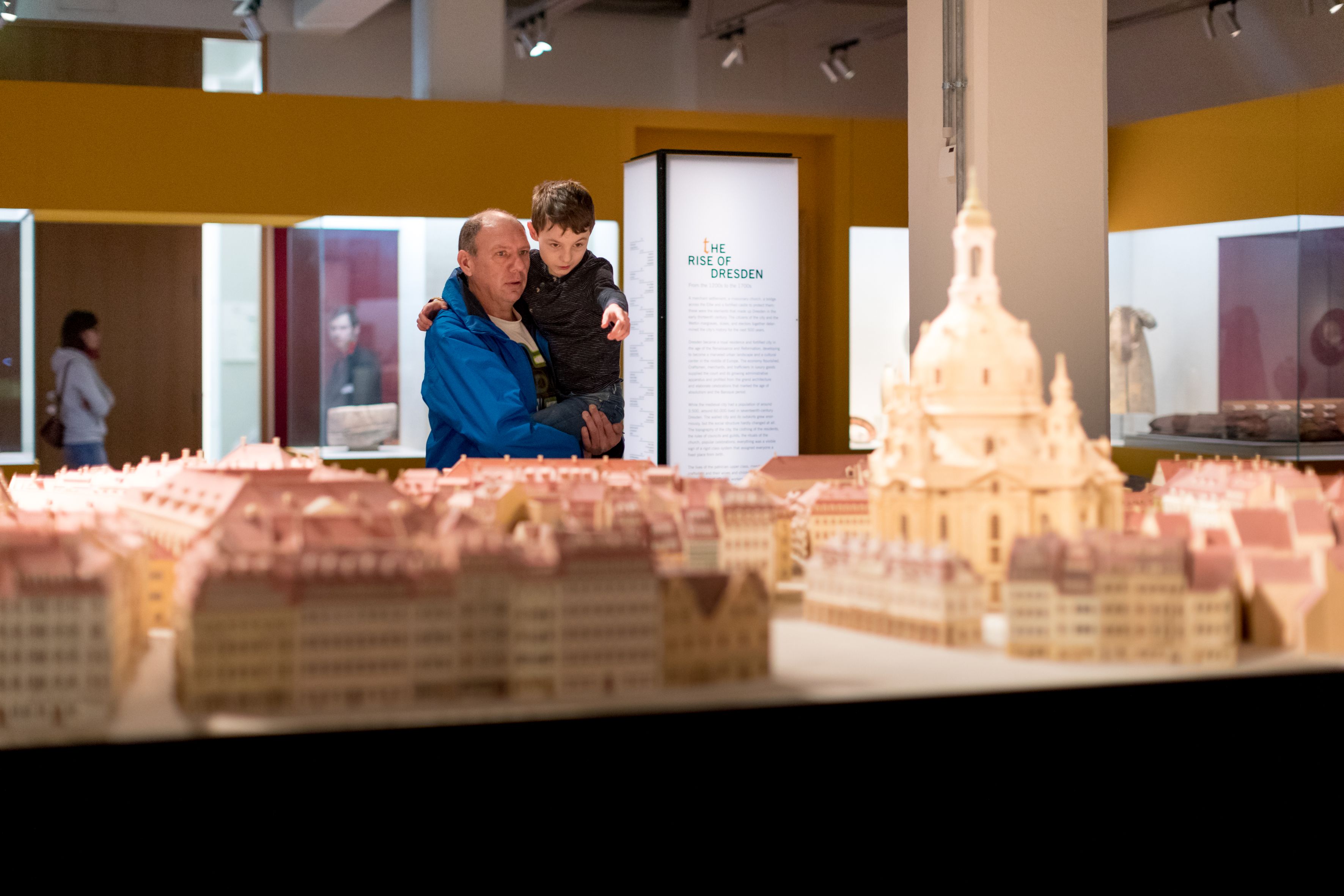 KI generiert: Ein Erwachsener und ein Kind betrachten ein Modell der Stadt Dresden in einem Museum. Im Hintergrund ist eine unscharfe Informationstafel mit der Aufschrift "The Rise of Dresden" zu sehen.