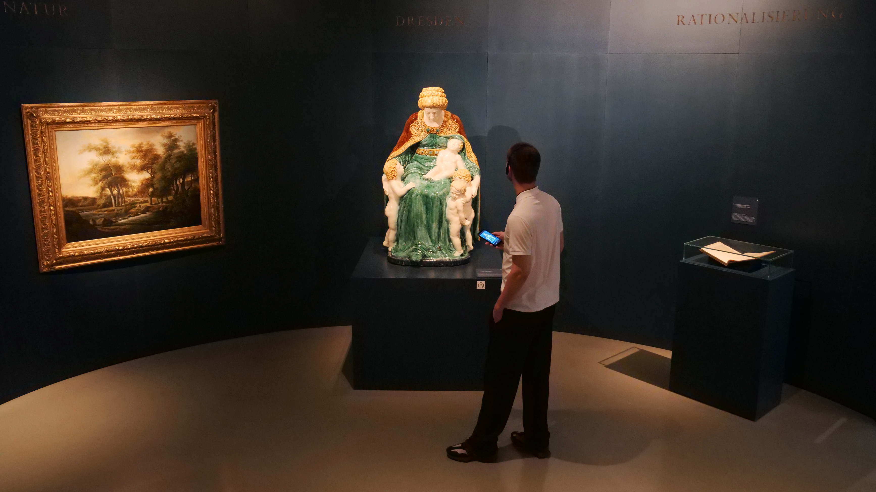 KI generiert: Ein Mann betrachtet in einem Museumsraum eine farbenfrohe Skulptur, die auf einem Podest ausgestellt ist. An der Wand hängt ein gerahmtes Gemälde.