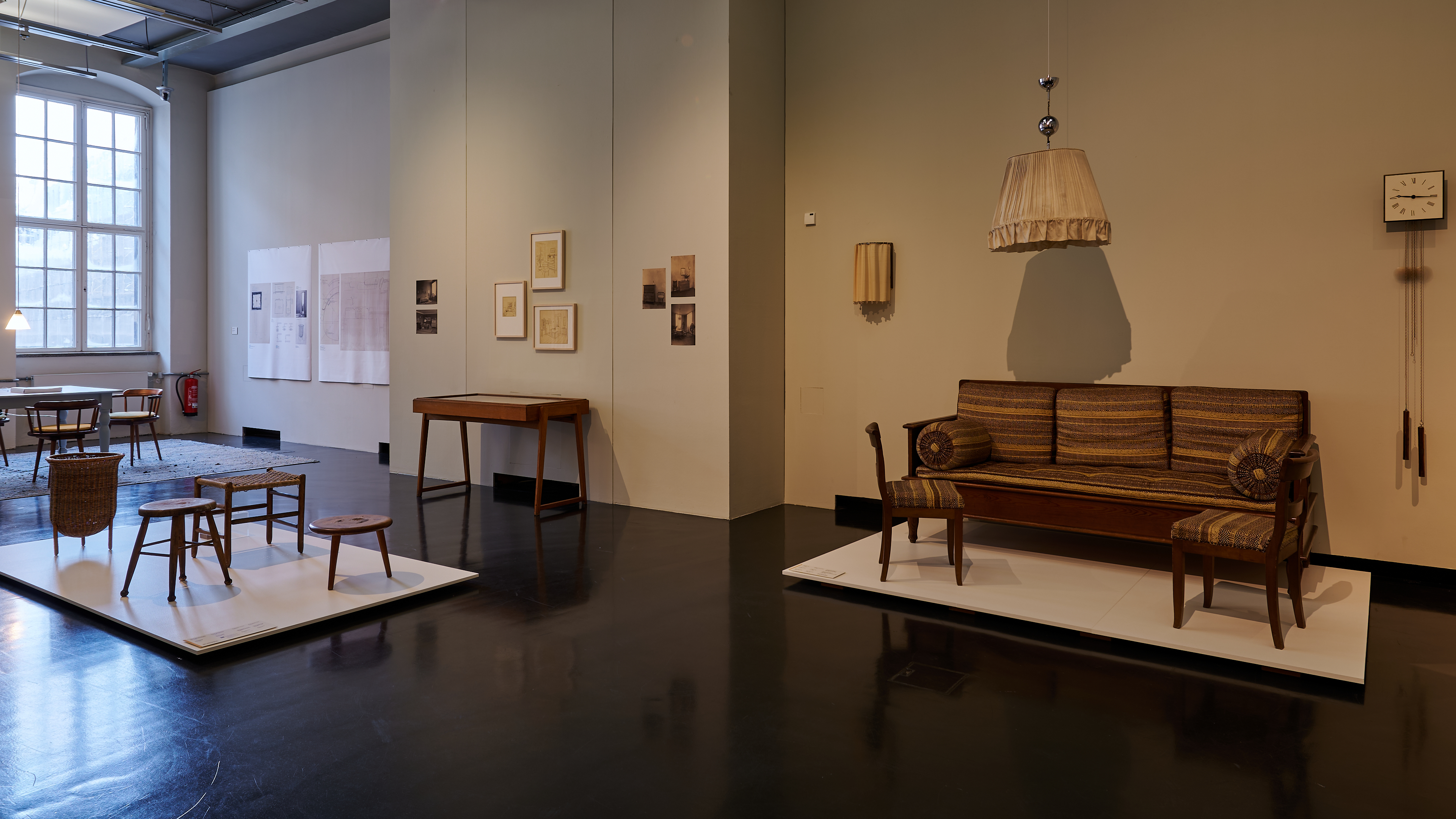 KI generiert: Das Bild zeigt eine Ausstellung von Möbelstücken in einem Museum oder einer Galerie, mit verschiedenen Sitzgelegenheiten und Tischen im Stil des frühen bis mittleren 20. Jahrhunderts. Die Anordnung ist auf hellen Podesten platziert, um die Designs zu betonen, und an den Wänden hängen begleitende Fotografien und Dokumente.