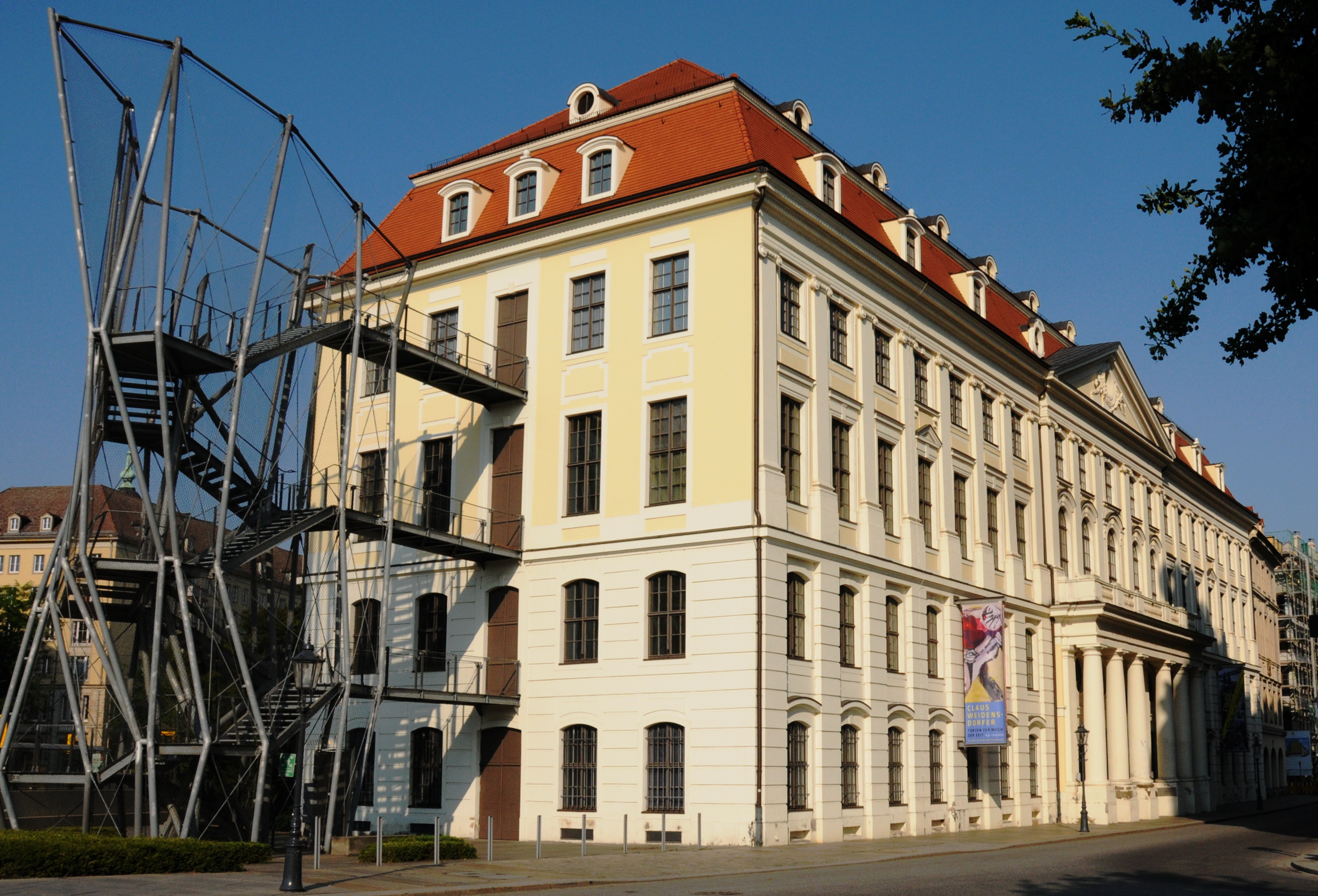 KI generiert: Das Bild zeigt ein großes, mehrstöckiges Gebäude mit einem markanten roten Dach und traditionellen architektonischen Merkmalen. Auf der linken Seite des Gebäudes befindet sich eine moderne Außentreppe aus Metall.