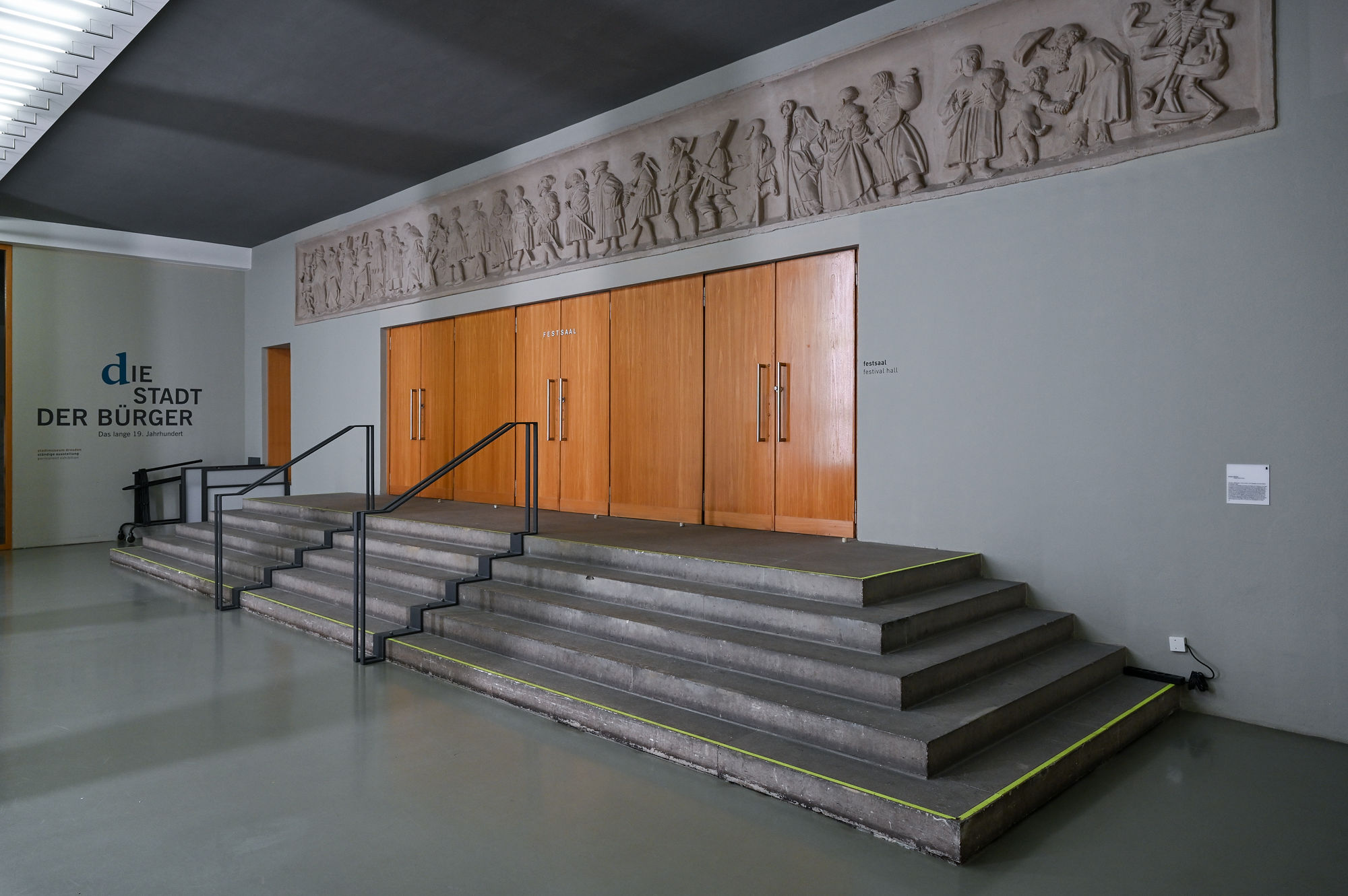 KI generiert: Das Bild zeigt eine Lobby oder einen Eingangsbereich mit Betonstufen, die zu Holz-Doppeltüren führen. Oberhalb der Türen befindet sich ein Relief mit geschnitzten Figuren, und an der linken Wand steht der Schriftzug "Die Stadt der Bürger".