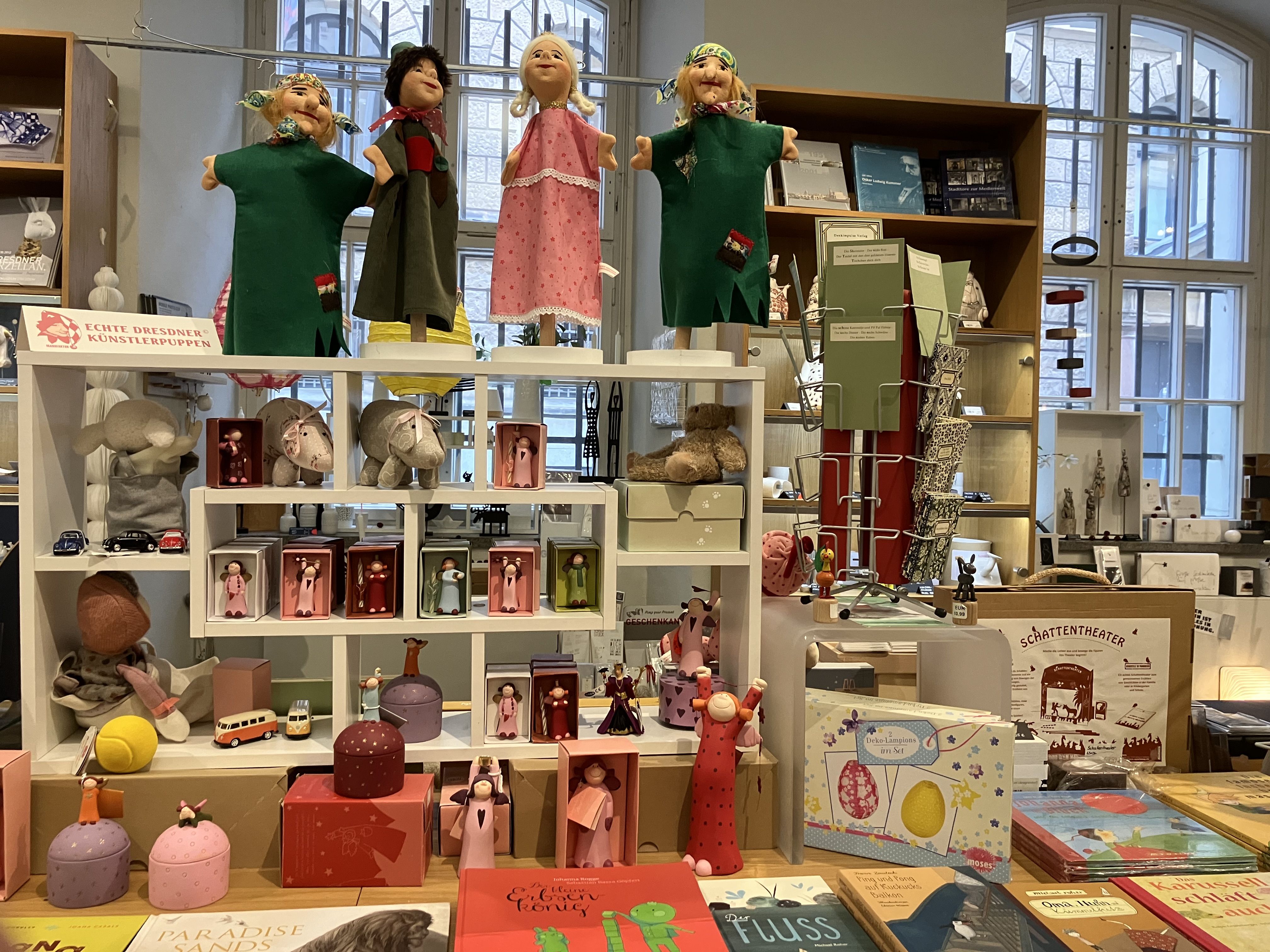 KI generiert: Das Bild zeigt eine bunte Ausstellung von Kunst- und Spielzeuggütern, darunter handgefertigte Puppen auf einem Regal. Im Vordergrund sind verschiedene Bücher und Dekorationsartikel sichtbar.