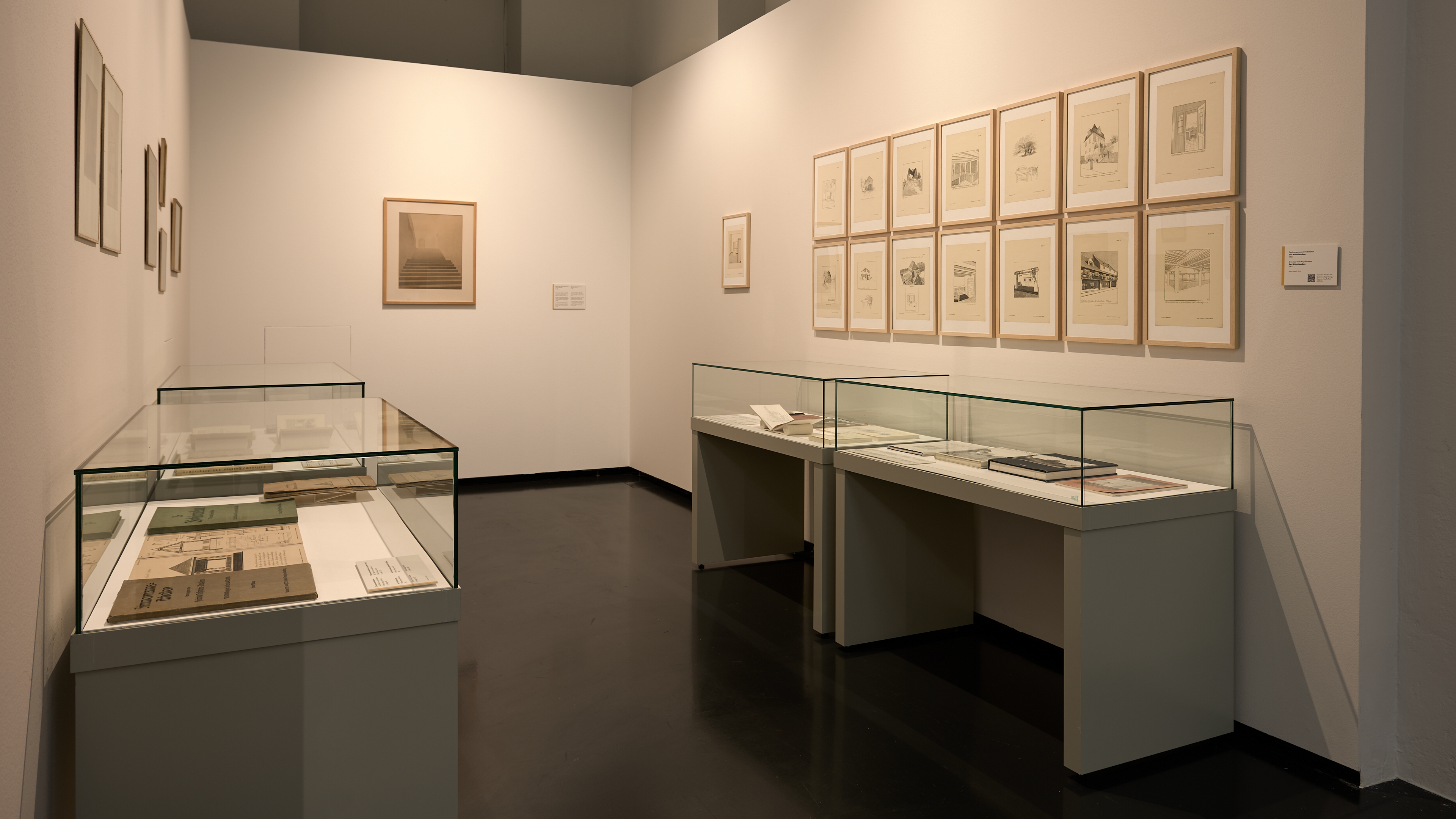 KI generiert: Das Bild zeigt einen Ausstellungsraum in einem Museum oder einer Galerie mit gerahmten Zeichnungen an den Wänden und mehreren Vitrinen, die Dokumente oder Kunstwerke präsentieren. Der Raum wirkt gut beleuchtet und ordentlich arrangiert.