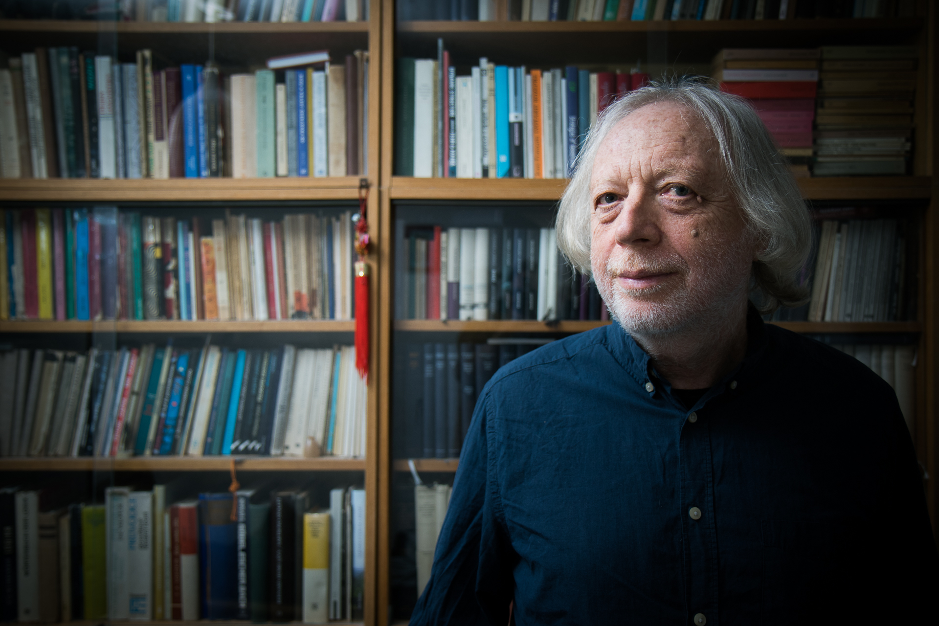 KI generiert: Ein Mann mit grauen Haaren steht vor einem großen Bücherregal und schaut leicht lächelnd in die Kamera. Der Hintergrund ist mit einer Vielzahl von Büchern gefüllt, was auf eine studierte oder akademische Umgebung hindeutet.