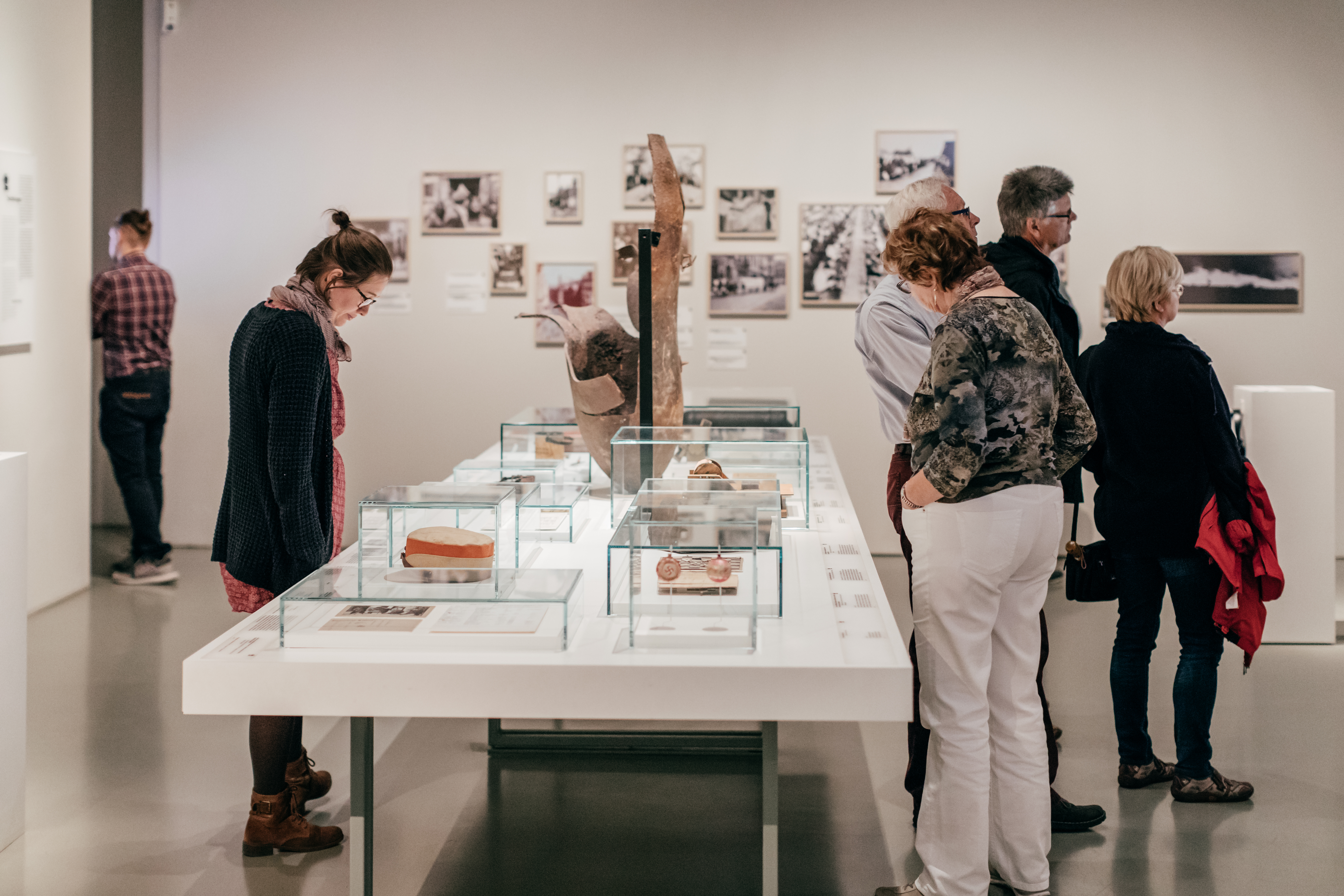KI generiert: Das Bild zeigt mehrere Personen, die in einem Museum oder einer Ausstellungssaal Exponate in Glasvitrinen betrachten. An den Wänden im Hintergrund sind weitere Bilder und Fotografien zu sehen.