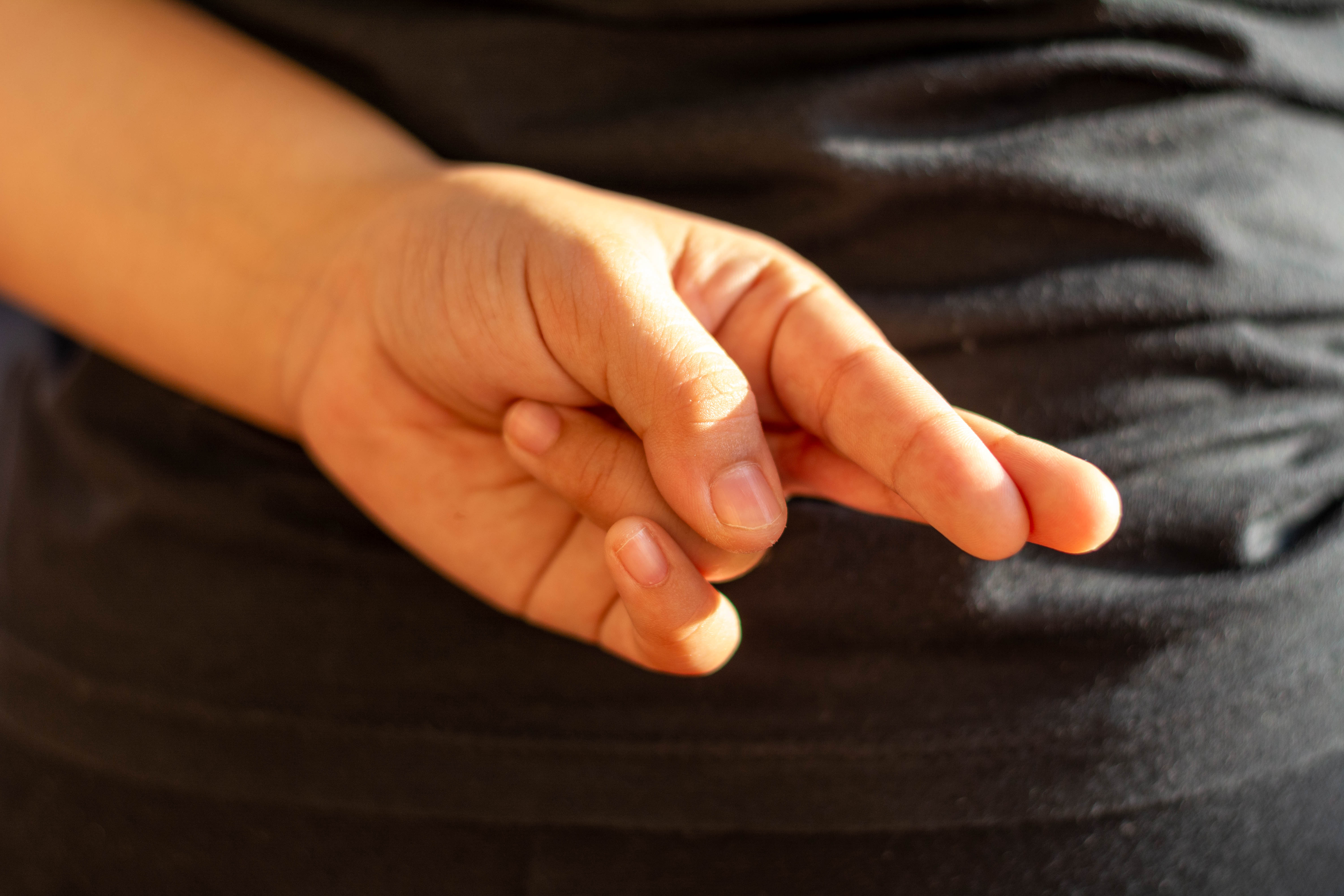 KI generiert: Das Bild zeigt eine Hand mit gekreuzten Fingern. Die Person trägt dunkle Kleidung.