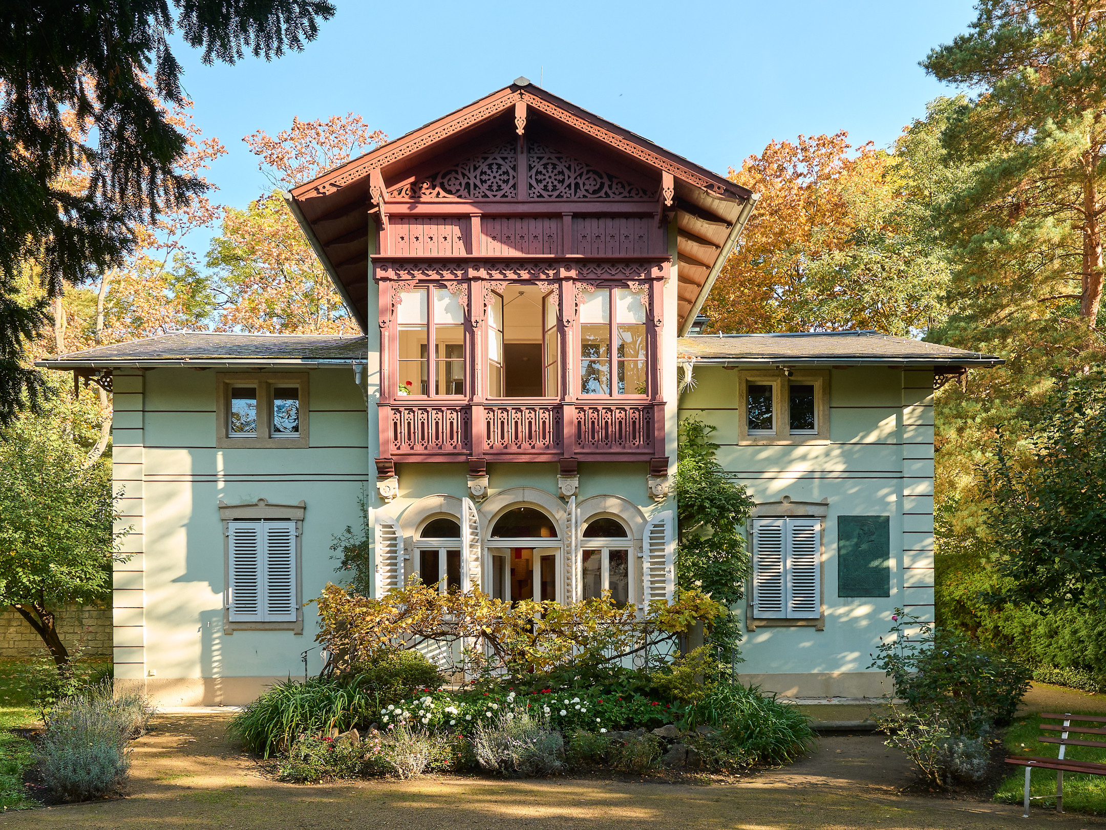 KI generiert: Das Bild zeigt ein zweistöckiges Gebäude mit einer markanten, reich verzierten Holzveranda im Obergeschoss. Es ist von üppigem Grün und einem gepflegten Garten umgeben.