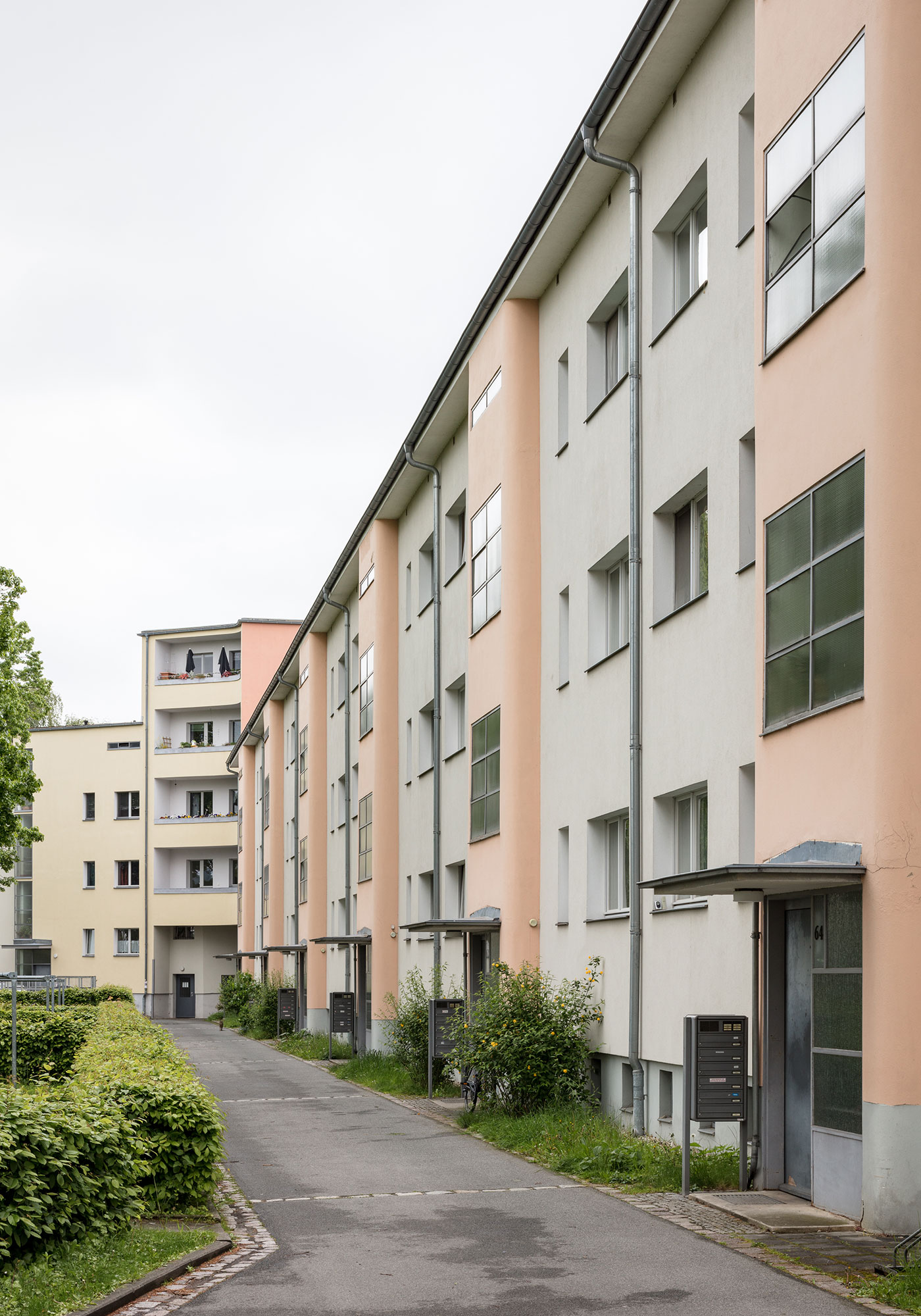 KI generiert: Das Bild zeigt eine Reihe von Mehrfamilienhäusern mit pastellfarbenen Fassaden entlang eines Gehwegs. Die Umgebung ist gepflegt mit Hecken und Grünflächen.