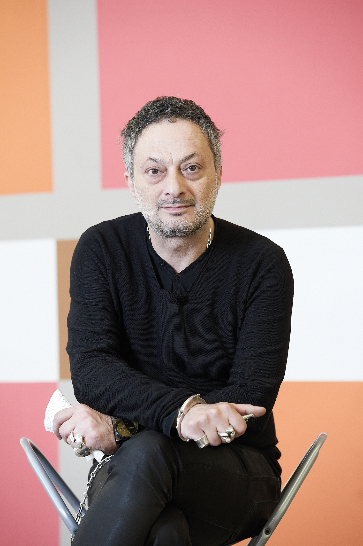 KI generiert: Das Bild zeigt eine Person, die auf einem Stuhl sitzt und in die Kamera blickt. Der Hintergrund ist in verschiedenen Farbfeldern gestaltet.