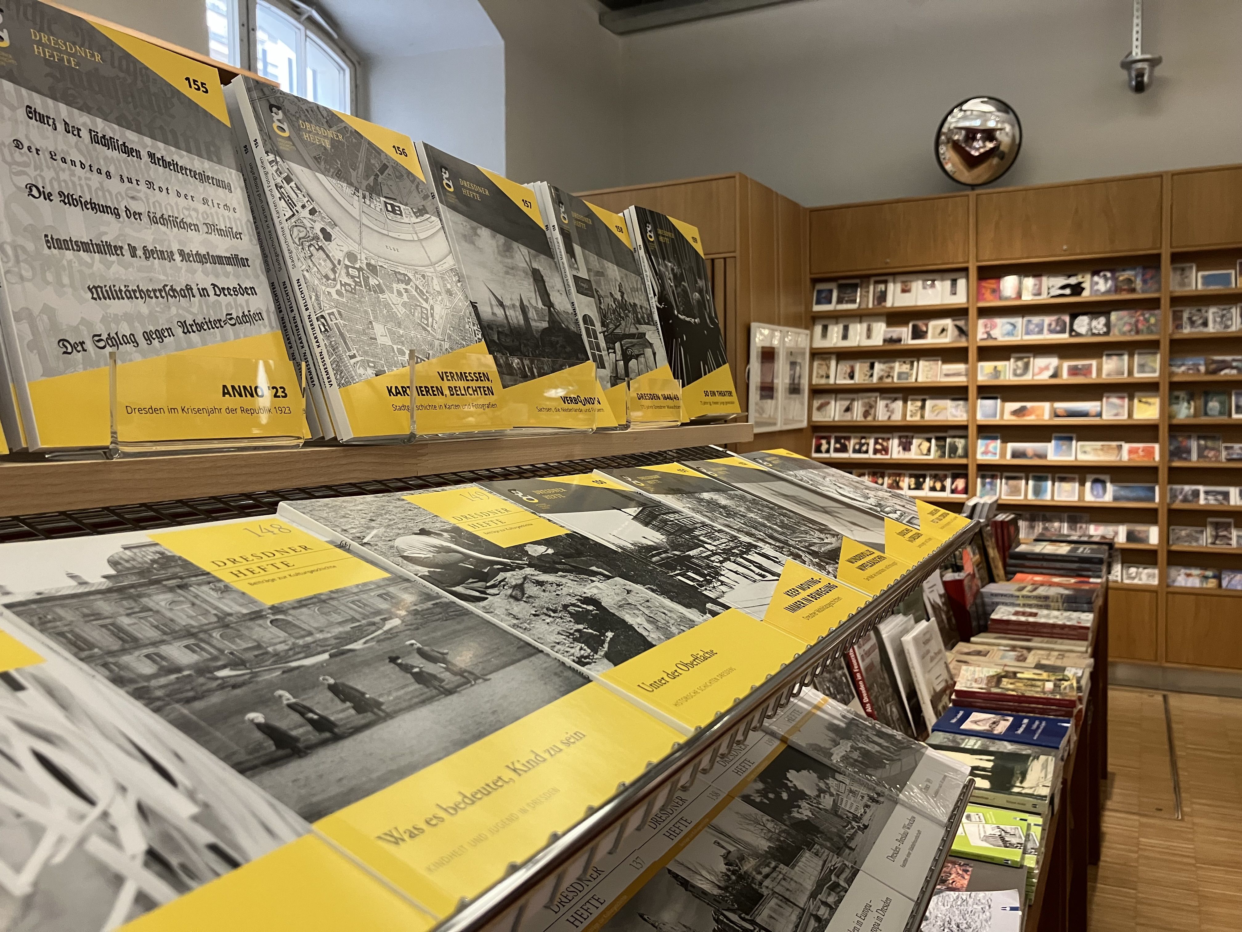 KI generiert: Das Bild zeigt ein Regal in einem Buchladen mit historischen Zeitschriften oder Broschüren, die auf Deutsch beschriftet sind. Im Hintergrund sind weitere Regale mit Büchern oder Postkarten zu sehen.