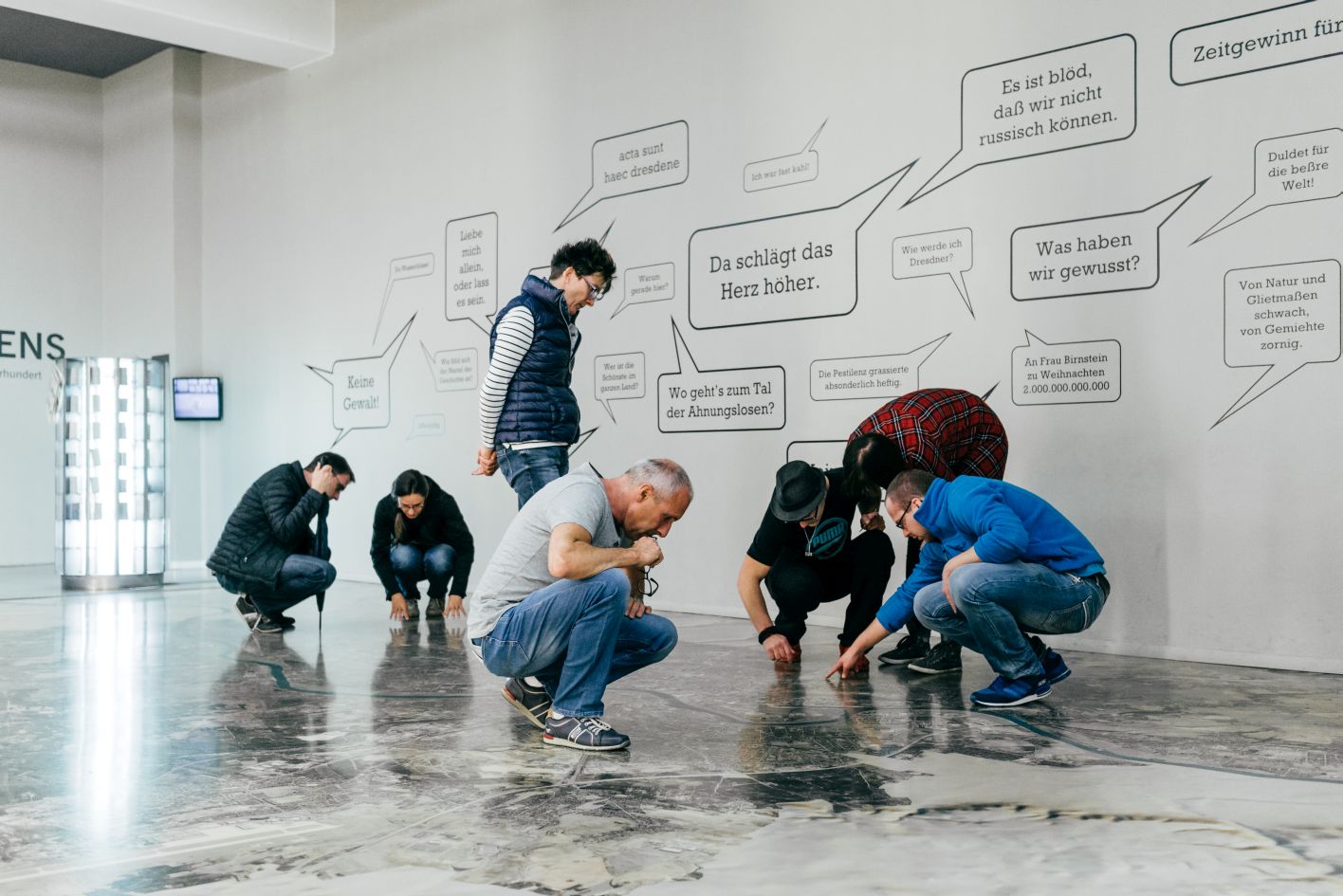 KI generiert: Das Bild zeigt eine Gruppe von Menschen, die sich auf einem Boden versammelt haben und diesen anscheinend untersuchen oder betrachten. Im Hintergrund sind zahlreiche Sprechblasen mit deutschen Texten an einer weißen Wand zu sehen.
