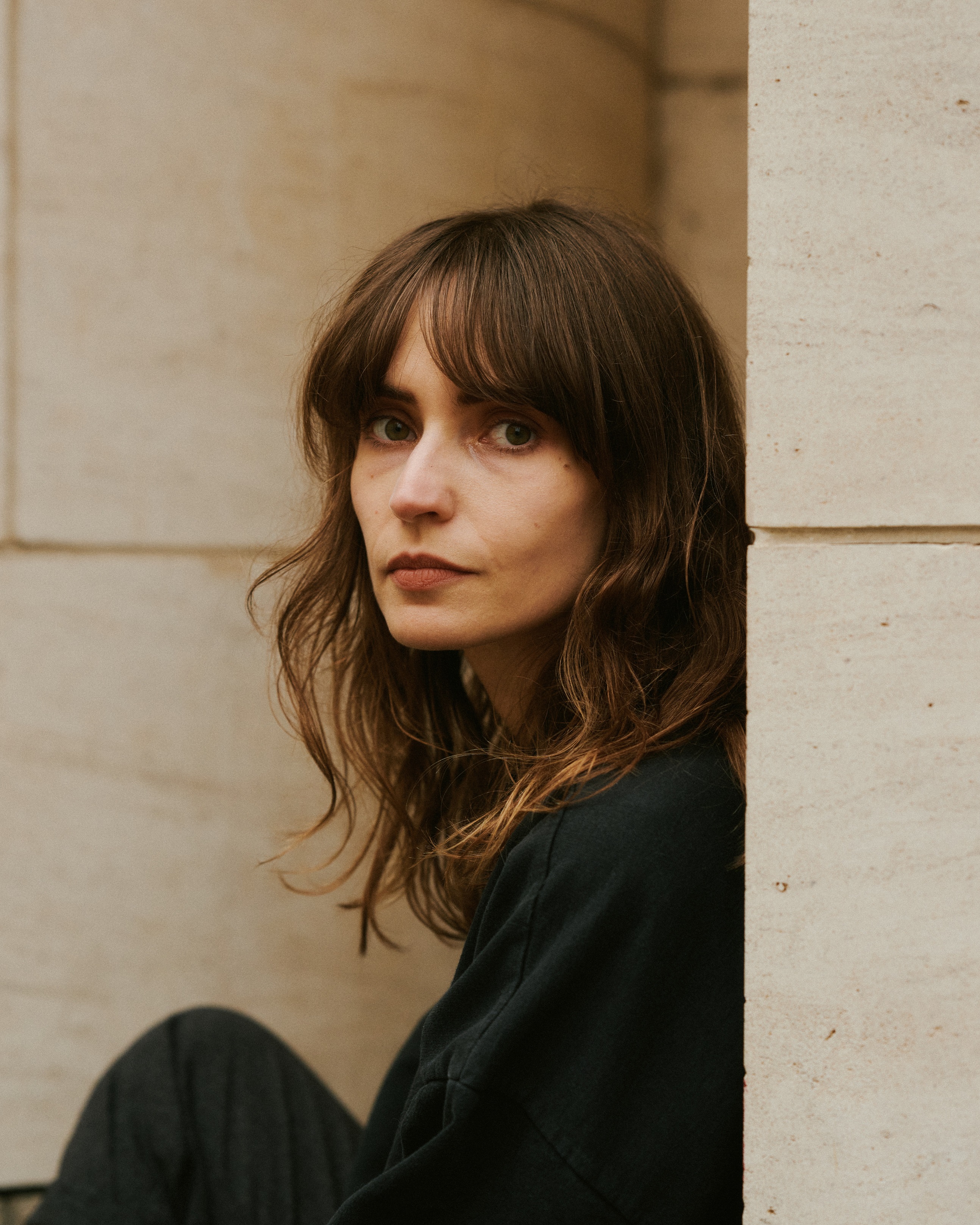 KI generiert: Das Bild zeigt eine Frau mit langen, braunen Haaren, die nachdenklich in die Kamera blickt. Sie lehnt an einer hellen Steinmauer.