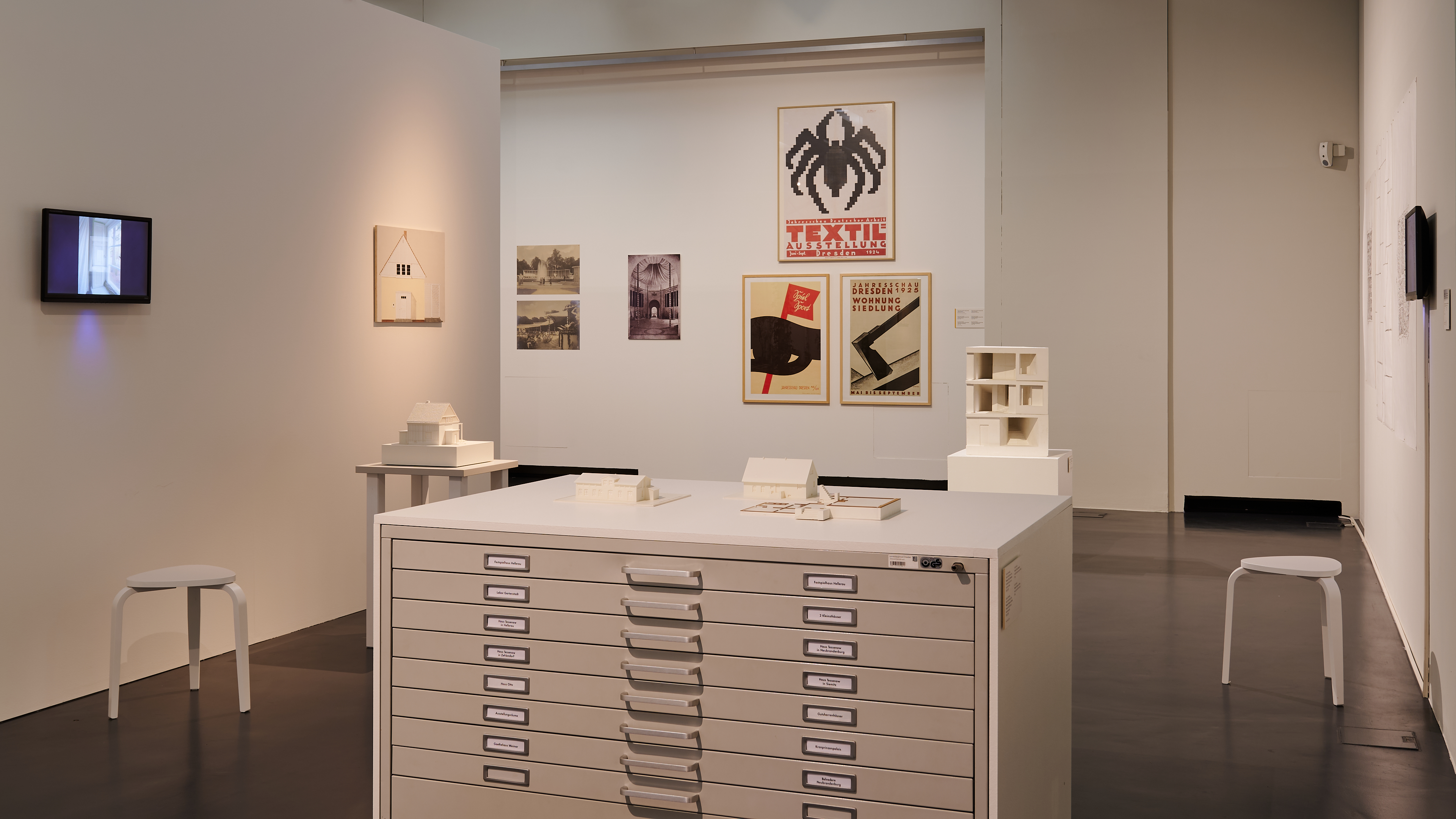 KI generiert: Das Bild zeigt einen Ausstellungsraum mit Zeichnungen an der Wand und Modellen auf Tischen. Im Vordergrund steht ein großer Schrank, auf dem kleine Architekturmodelle platziert sind.