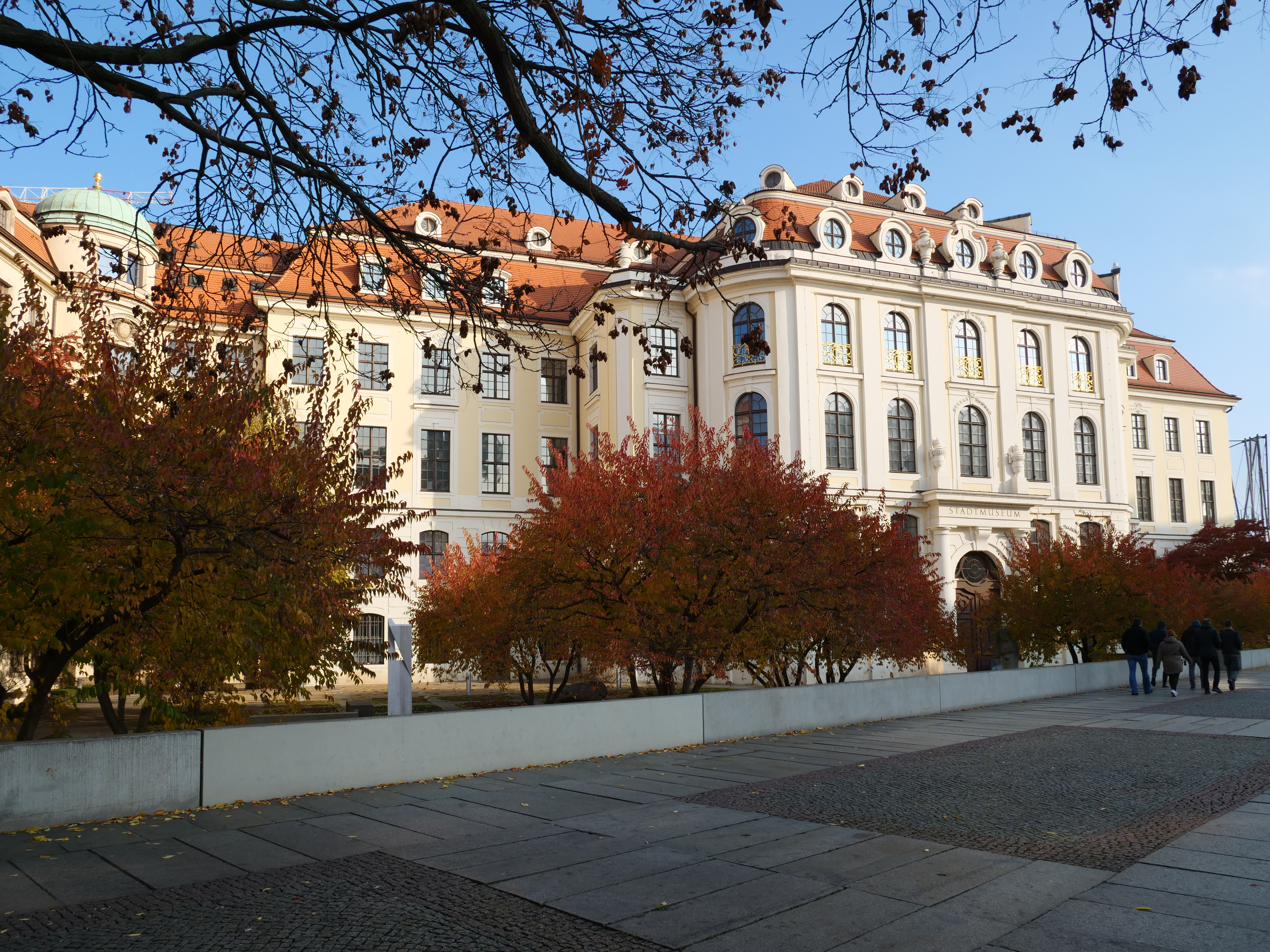 KI generiert: Das Bild zeigt ein historisches Gebäude mit einer eleganten Fassade und einem roten Ziegeldach, umgeben von Bäumen mit Herbstlaub. Vor dem Gebäude befinden sich ein Gehweg und einige Menschen, die vorbeigehen.