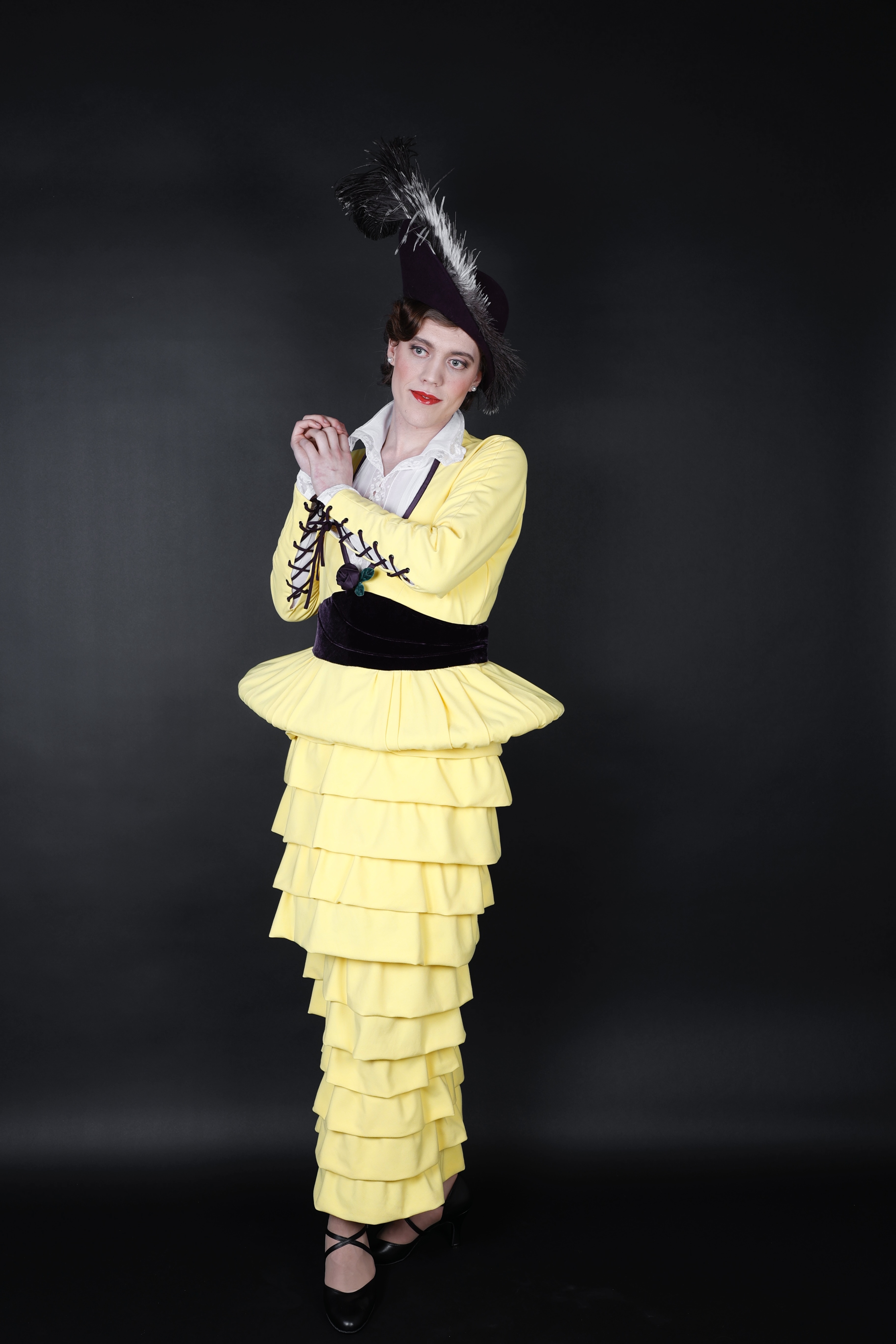 KI generiert: Das Bild zeigt eine Person in einem auffälligen, gelben Outfit mit mehreren Rüschen und einem dazu passenden Hut mit Feder. Der Hintergrund ist schlicht und dunkel, was den Fokus auf die Kleidung lenkt.