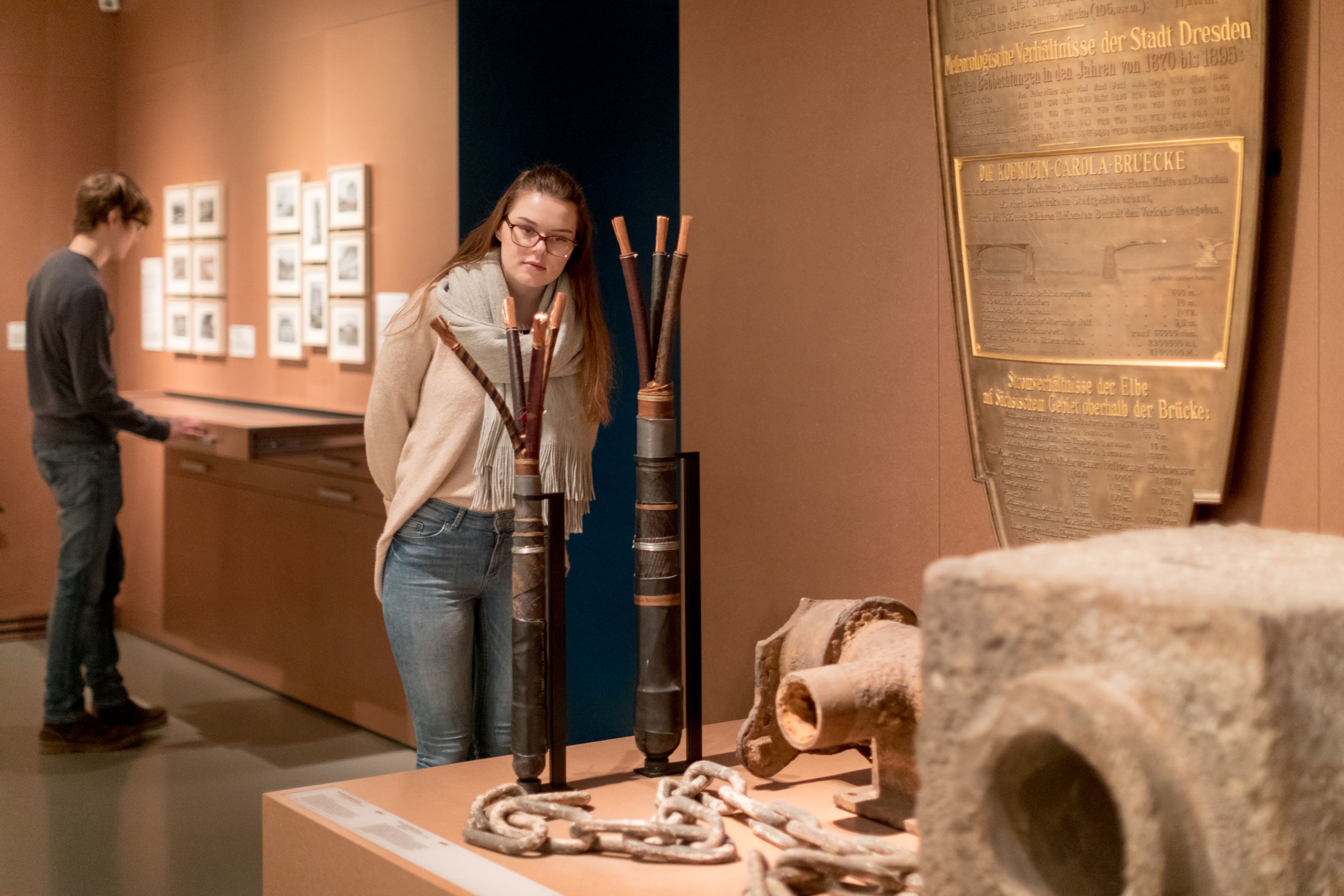 KI generiert: Das Bild zeigt zwei Personen in einem Museum, die sich verschiedene ausgestellte Objekte und Fotografien ansehen. Die Hauptperson im Vordergrund betrachtet interessiert einige historische Artefakte und eine Tafel.