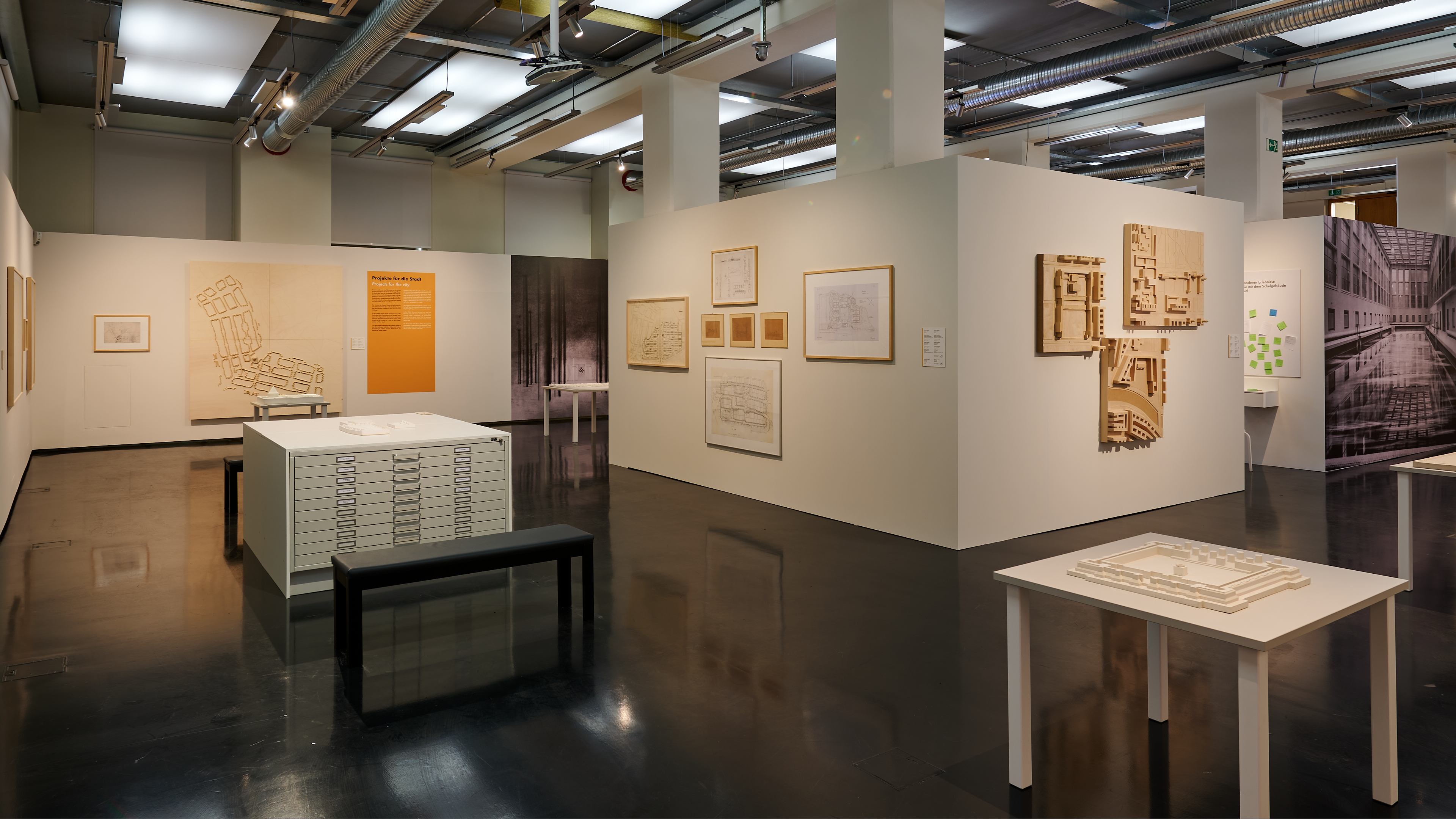 KI generiert: Das Bild zeigt eine Ausstellung in einem modernen, gut beleuchteten Galerieraum mit architektonischen Zeichnungen und Modellen. An den Wänden hängen Pläne und Entwürfe, während auf Tischen maßstabsgetreue Modelle stehen.