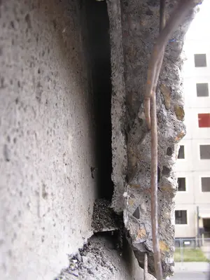 KI generiert: Das Bild zeigt einen beschädigten Betonabschnitt mit freiliegender Bewehrung und Rissen. Im Hintergrund ist ein mehrstöckiges Gebäude erkennbar.