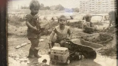 KI generiert: Das Bild zeigt Kinder, die in einer schlammigen Umgebung mit Spielzeugautos spielen. Im Hintergrund sind parkende Autos und Wohngebäude zu sehen.